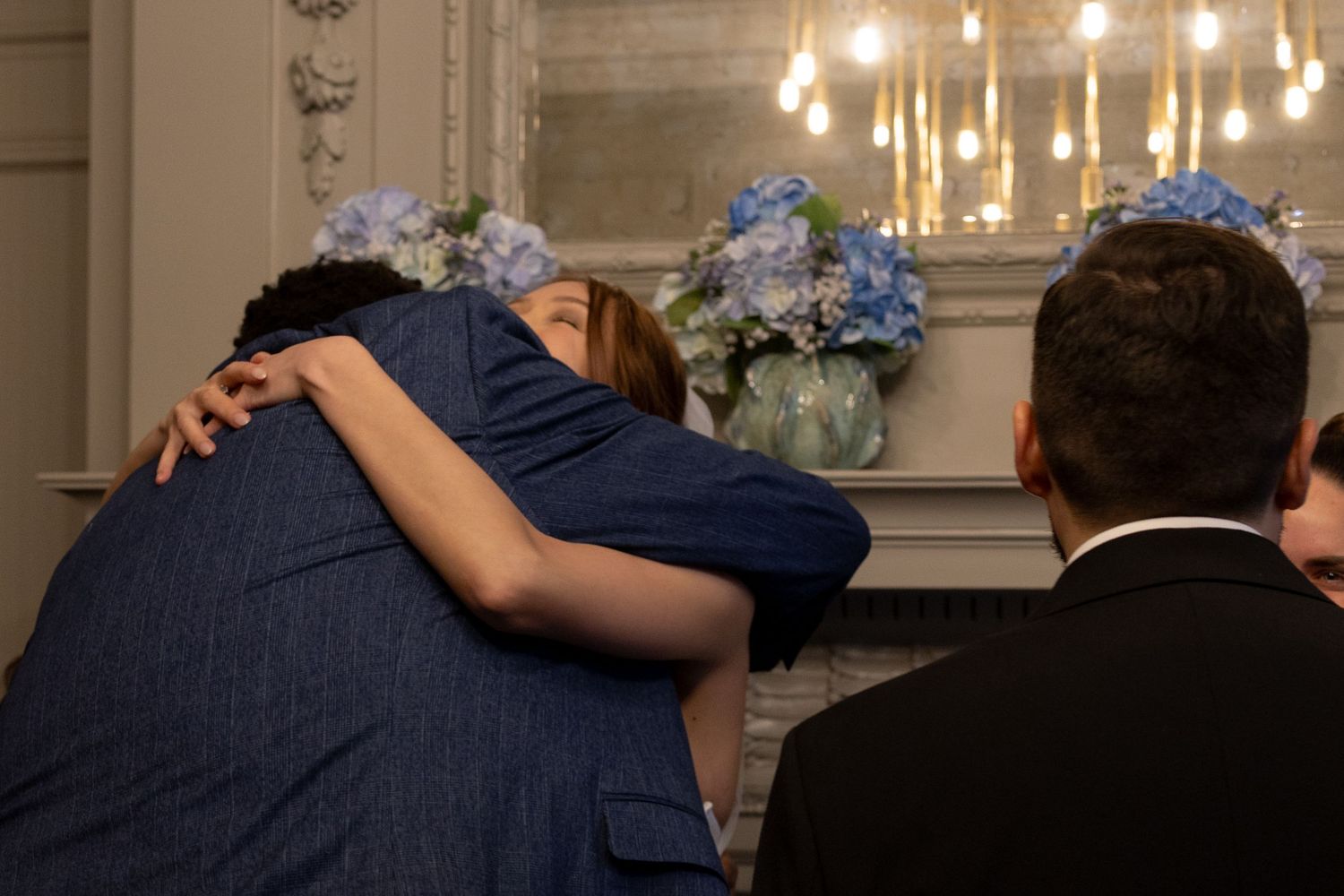 Wedding guest in blue dress sharing warm hug near floral arrangement of blue hydrangeas at reception.