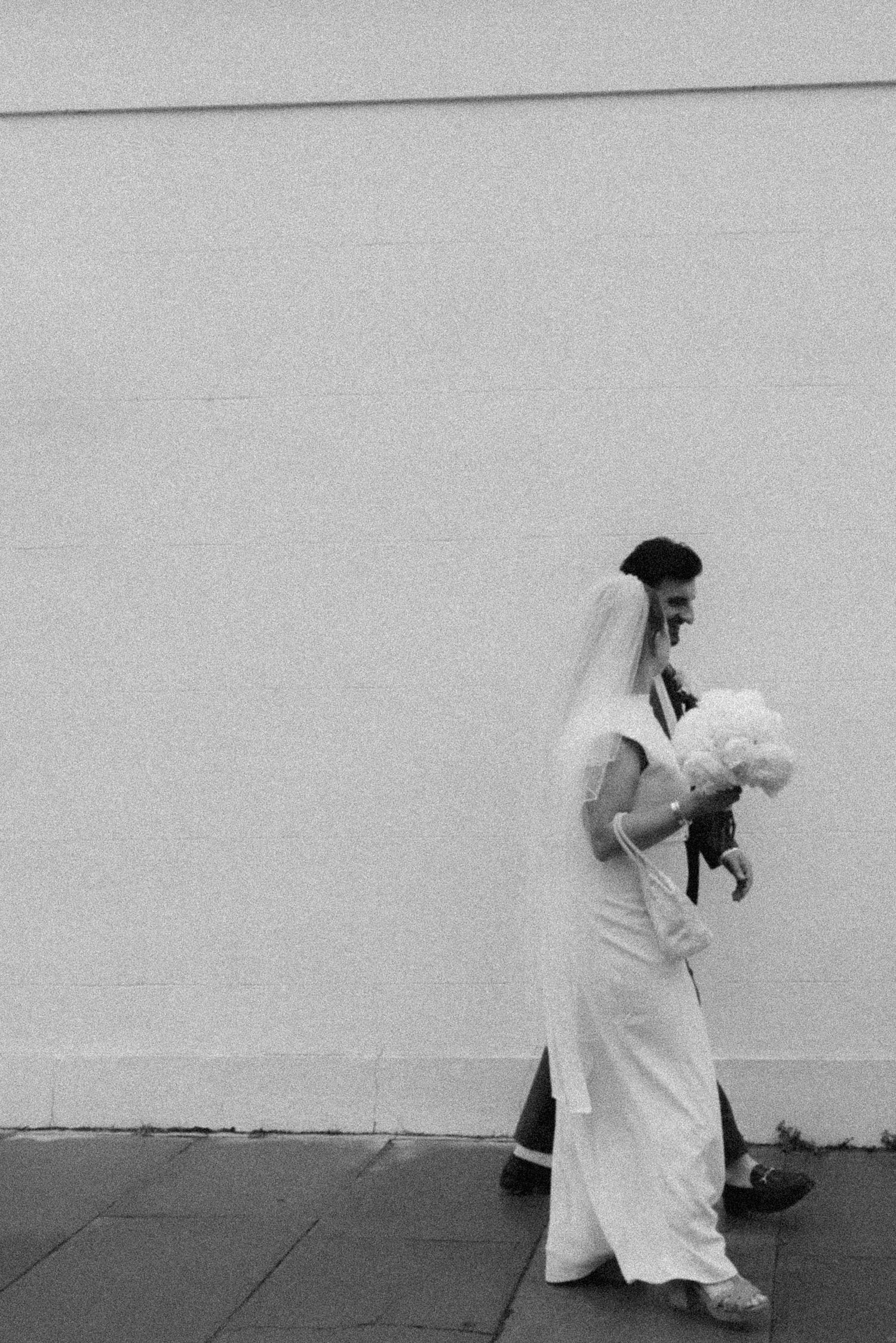 Black and white photo of a bride in white gown and veil walking with bouquet against a light wall.