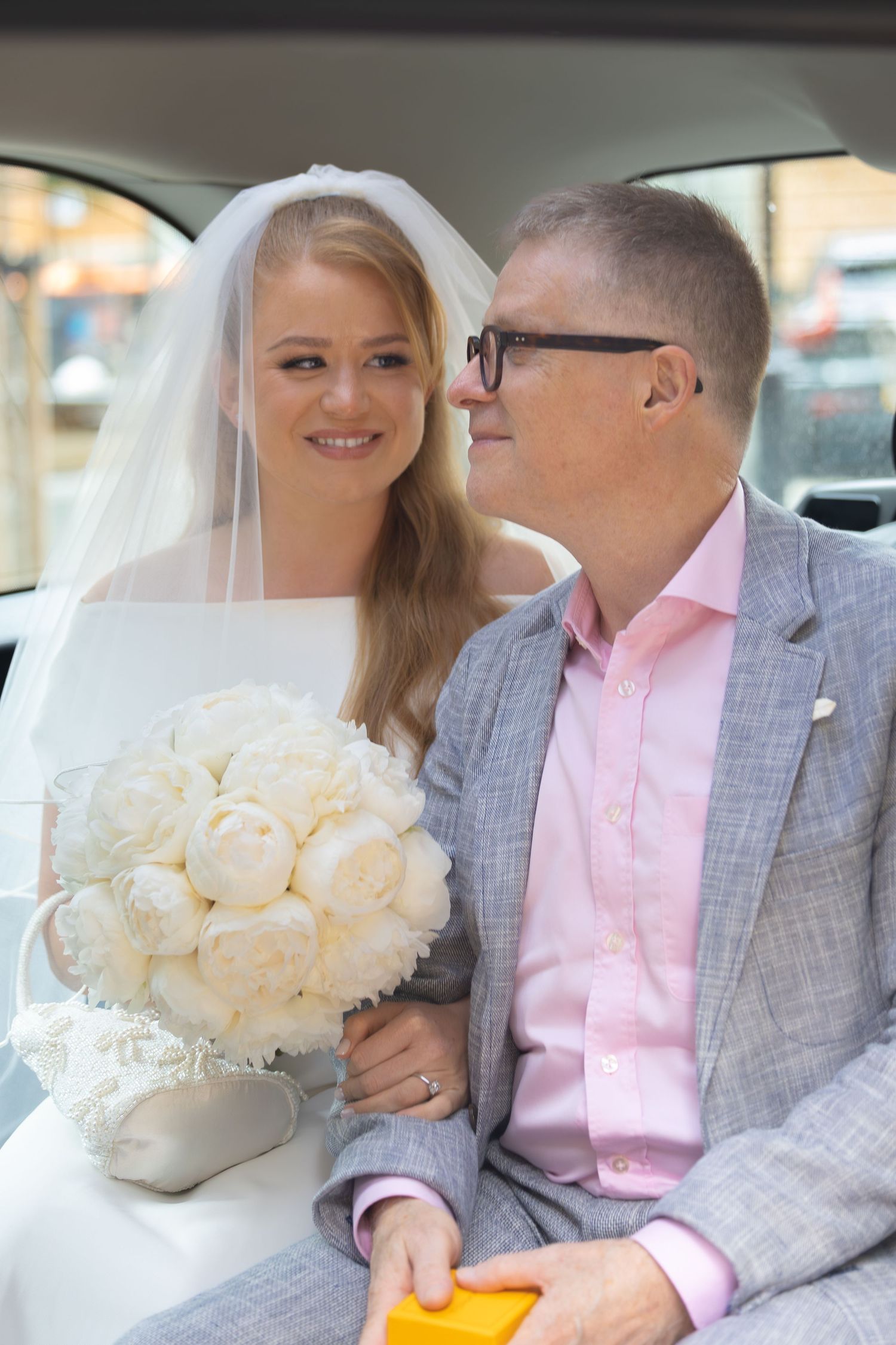 Bride in white veil holding bouquet of white peonies sits with groom in gray suit and pink shirt in car.