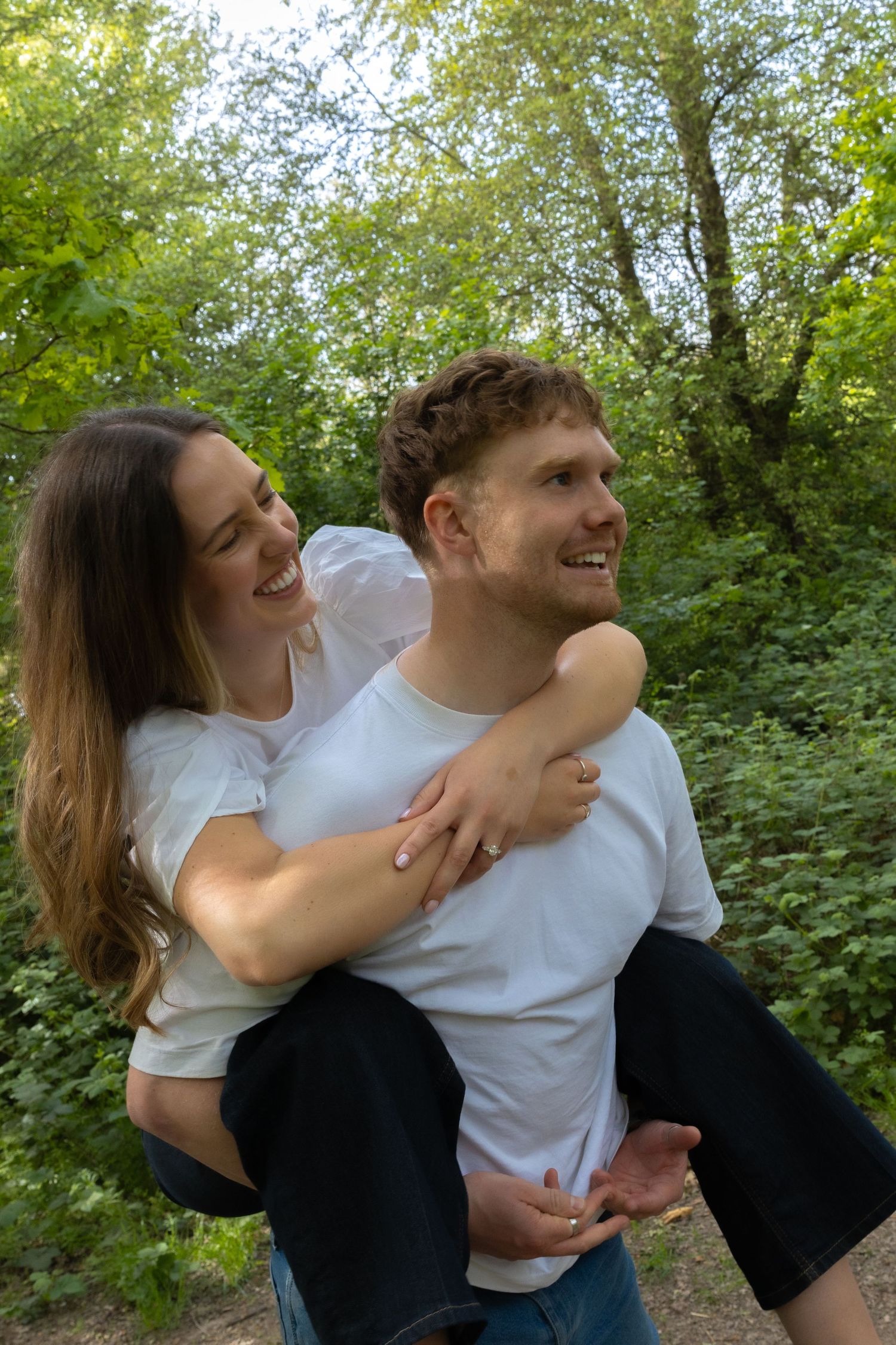 A happy couple smiling outdoors, man giving woman a piggyback ride in a lush green forest setting.