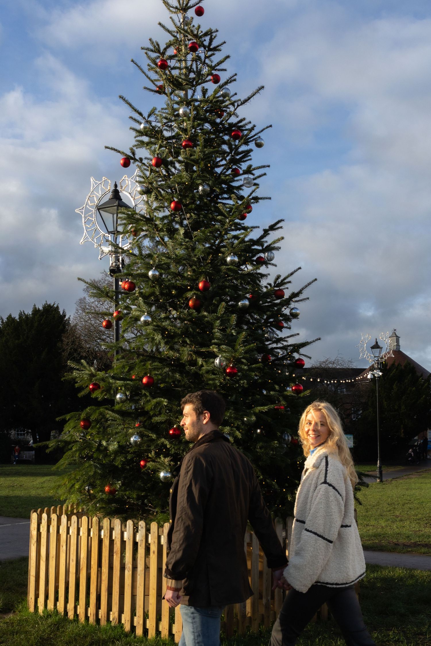 A couple stands near a decorated Christmas tree outdoors with a wooden fence in the background.