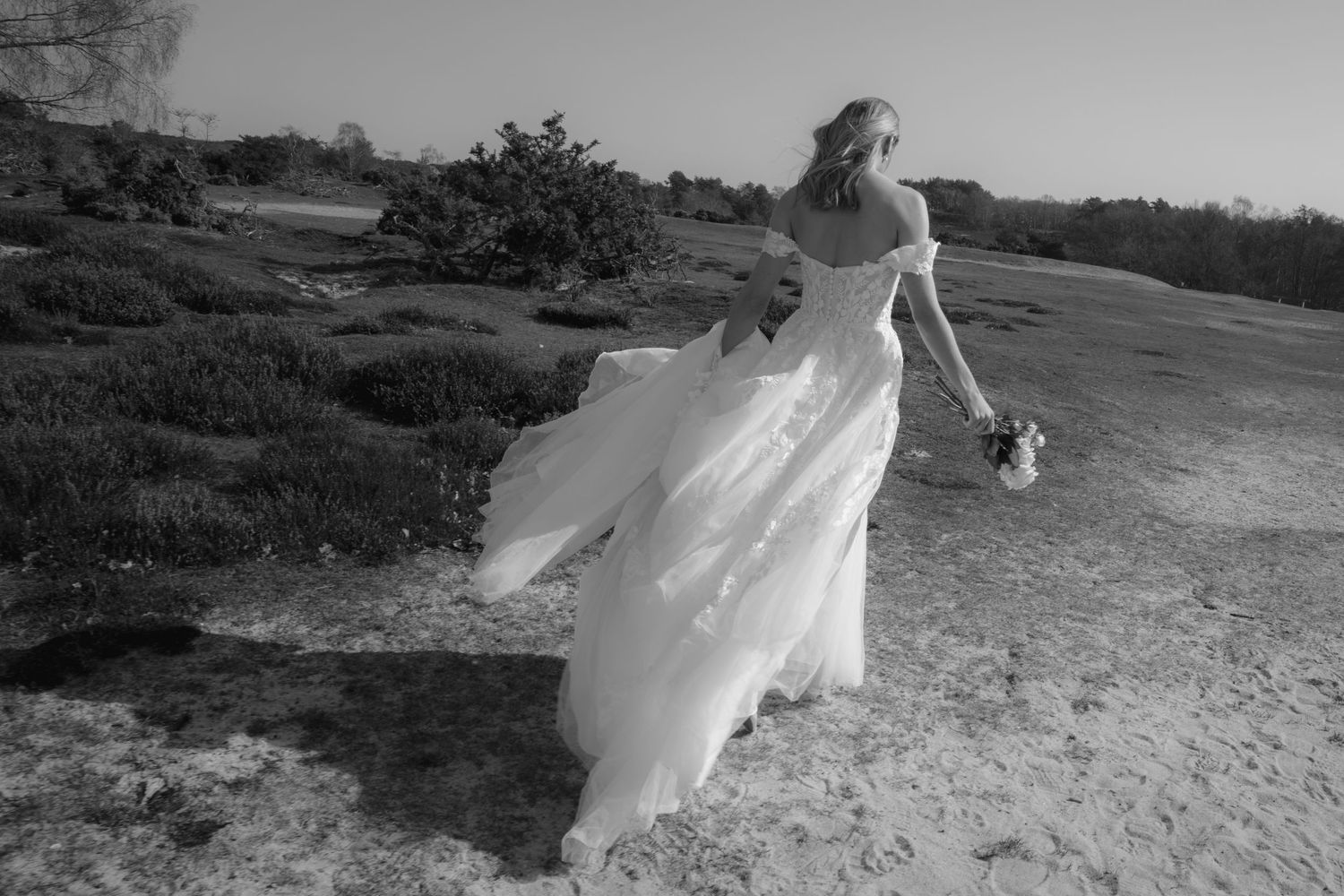 Black and white photo of a bride in a flowing wedding gown walking on sandy terrain with bouquet.