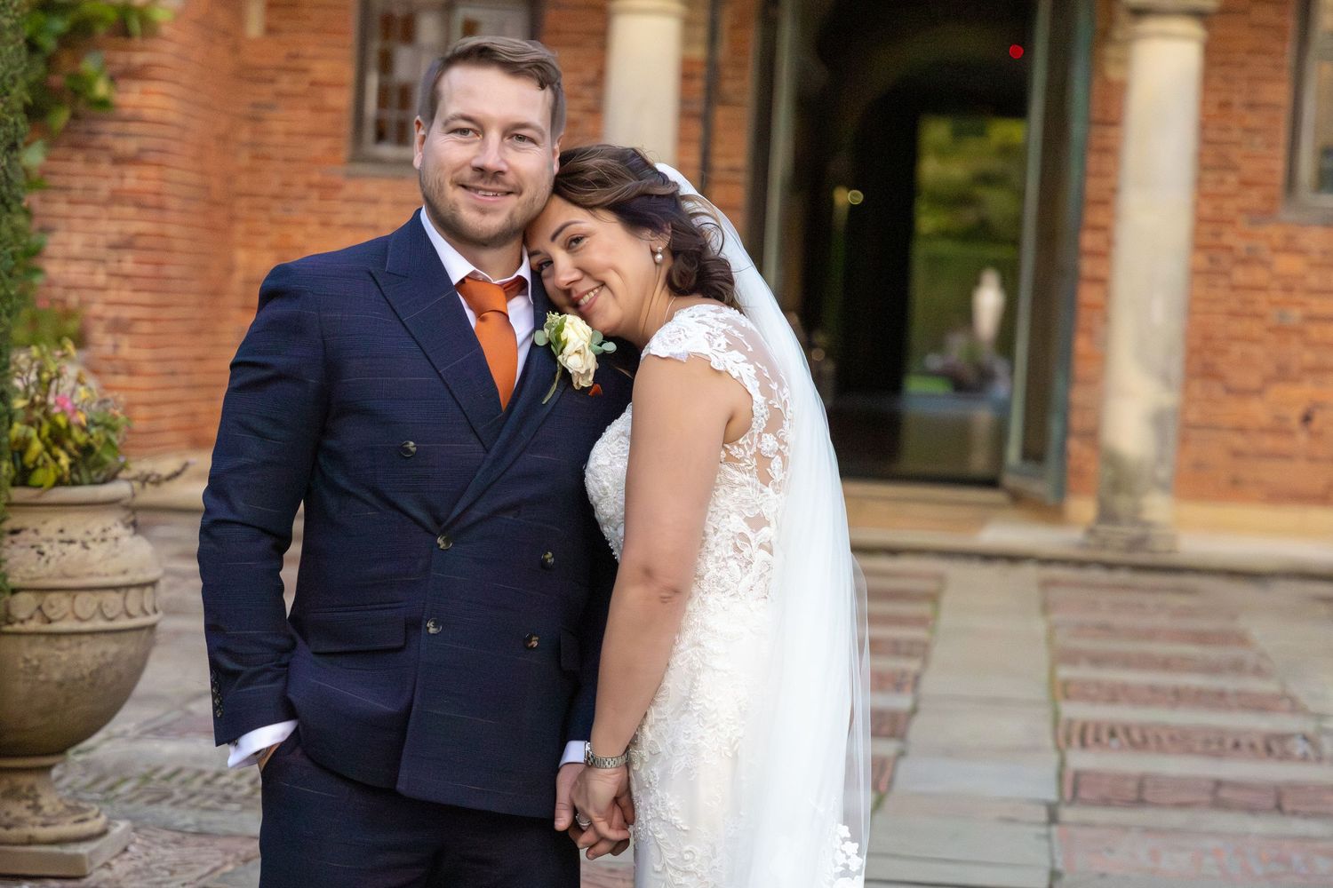 Couple in formal wedding attire posing in front of a brick building with arched windows.