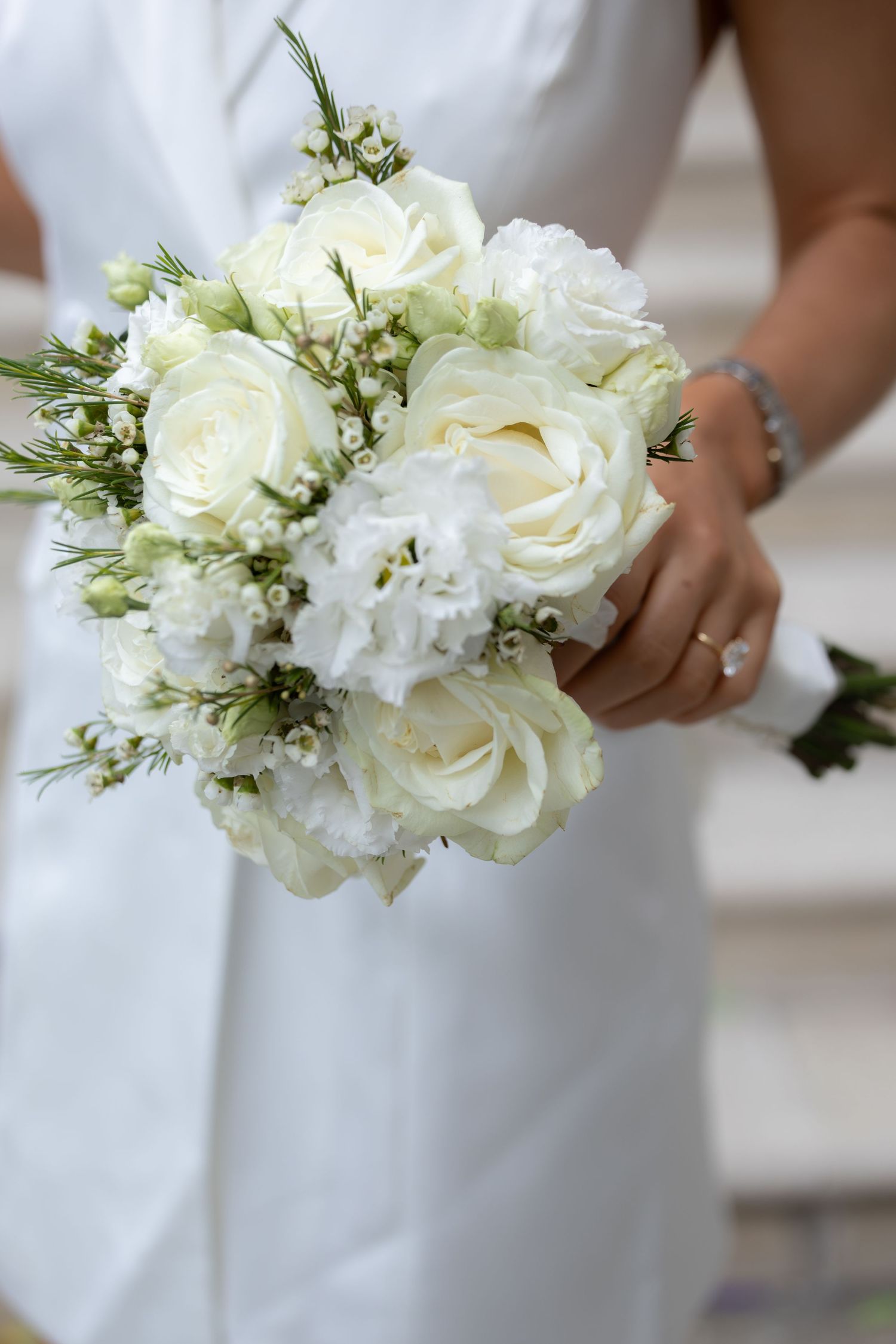 Bride in white dress holding elegant bridal bouquet of white roses and delicate flowers, wearing diamond ring.