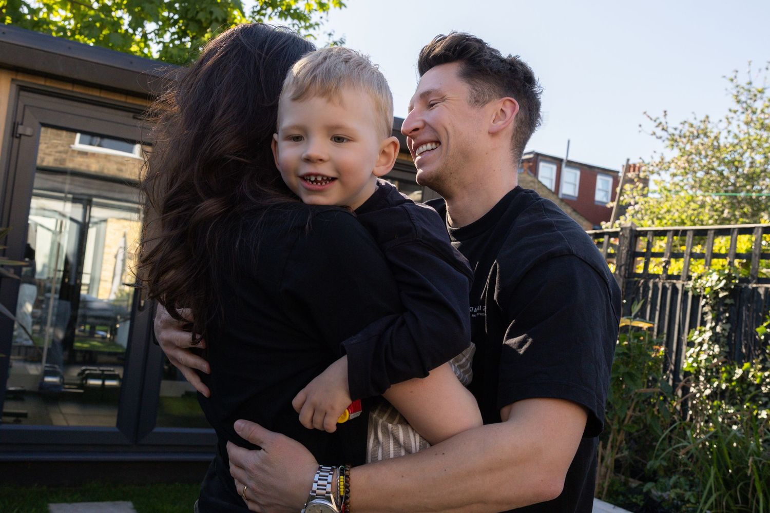 Laughing couple embraces a young smiling boy outdoors in a garden setting on a sunny day.