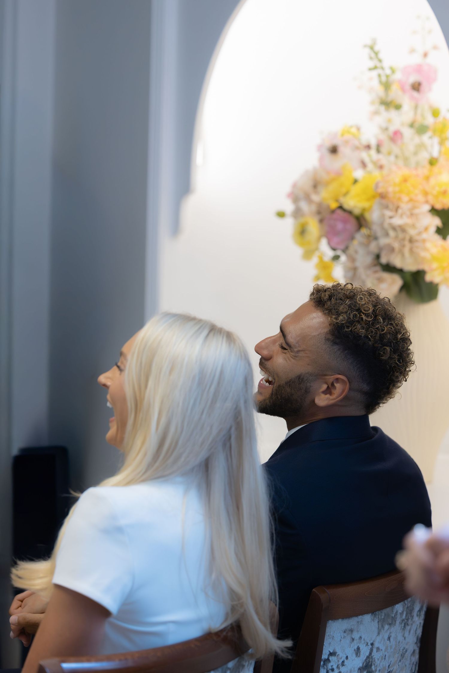 A laughing interracial couple sits together in a church, dressed formally, with floral decorations in the background.