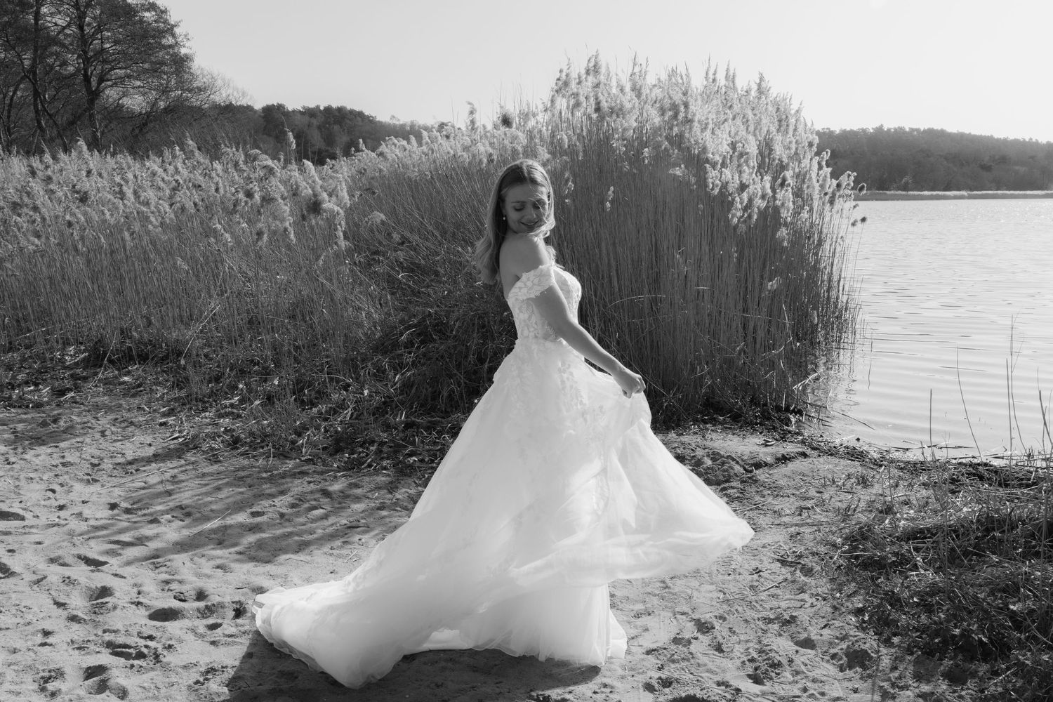 Black and white photo of a bride in a flowing wedding gown posing on a sandy shore near tall reeds and a lake.