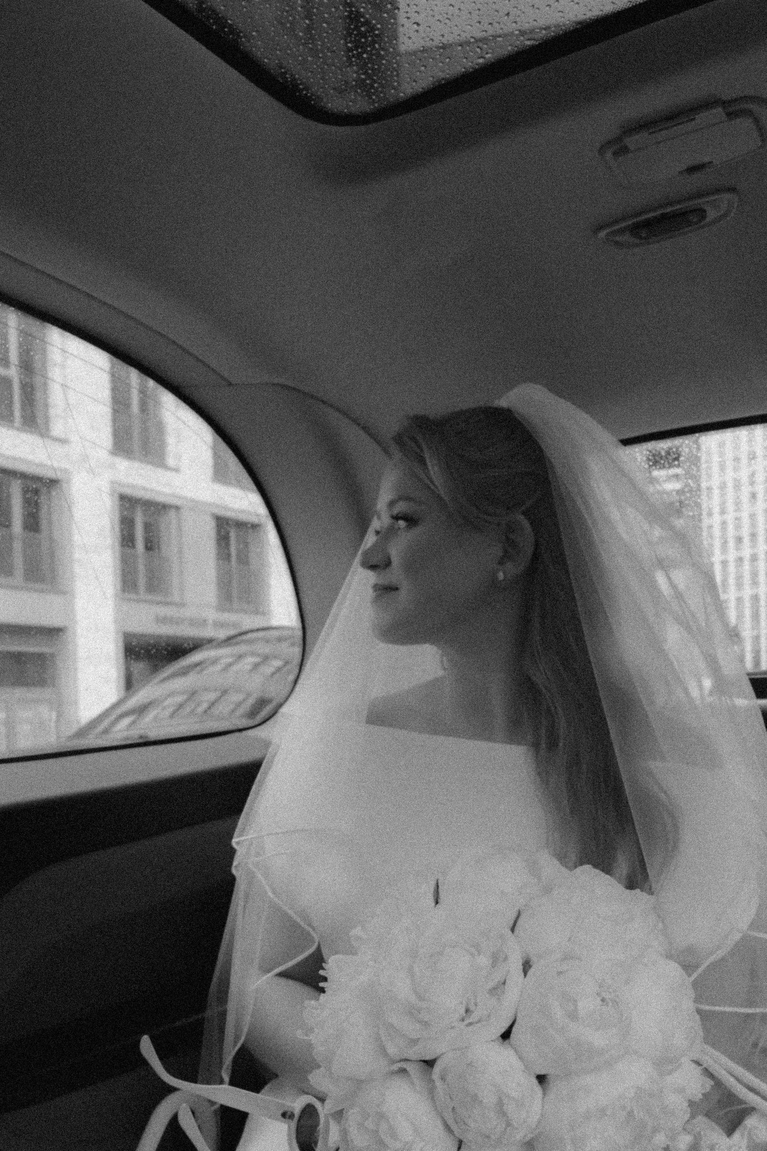 Elegant bride in veil holding white peony bouquet seated in car on wedding day.