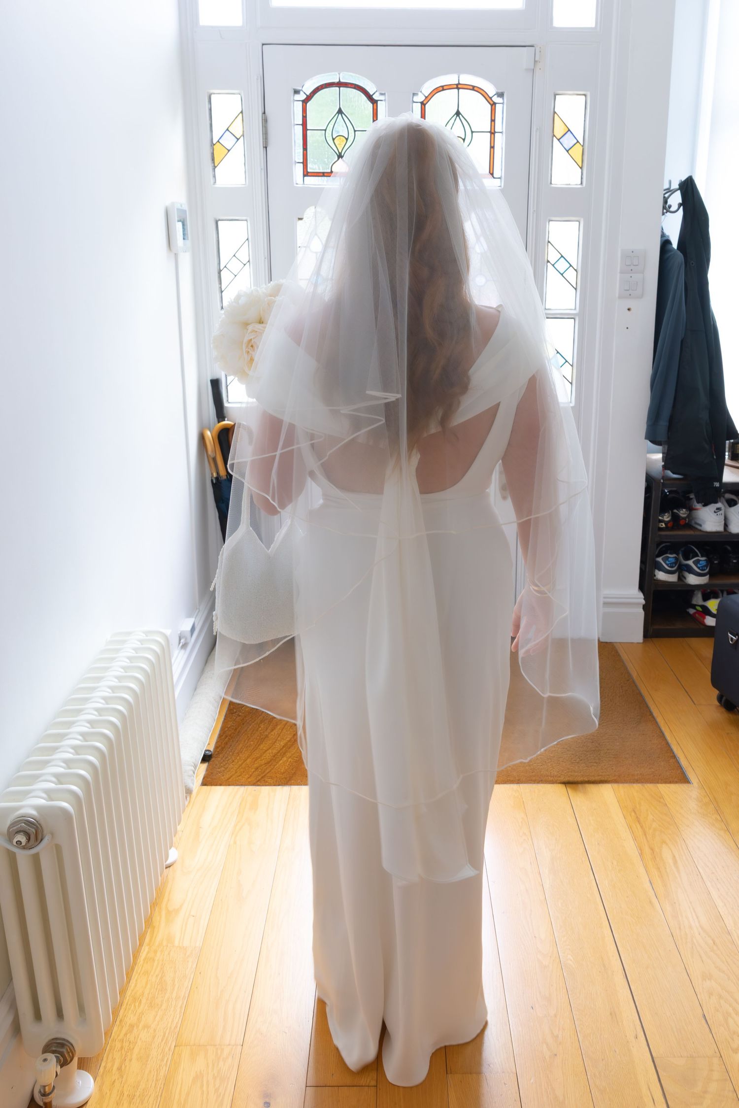 Bride in white wedding dress and veil stands at front door holding bouquet, viewed from behind.
