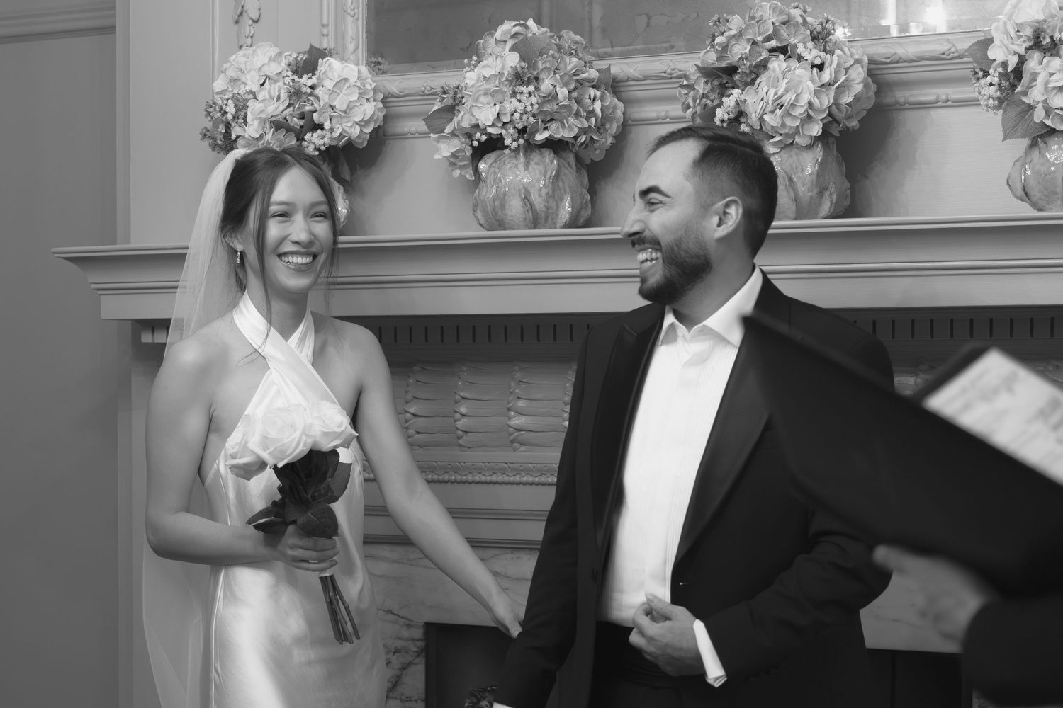 Black and white photo of a smiling bride holding bouquet and groom in tuxedo standing by fireplace at wedding ceremony.