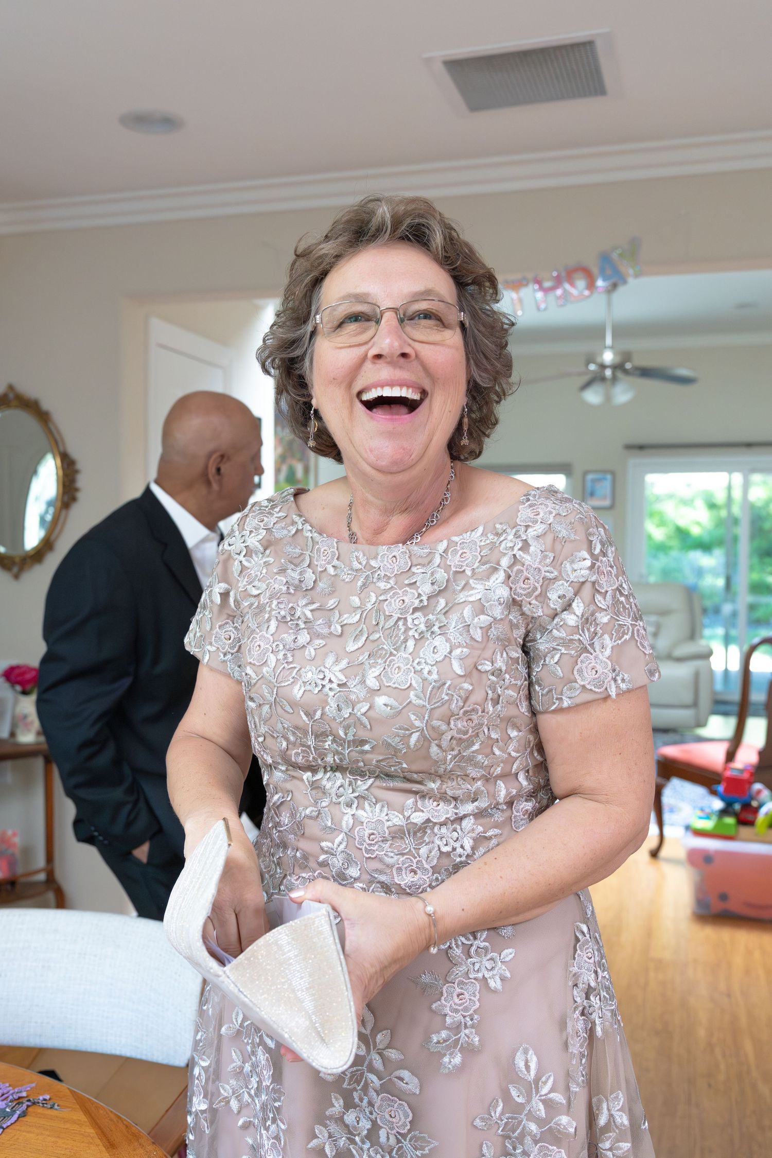 Elderly woman in elegant floral lace dress laughing joyfully at a wedding reception indoors.