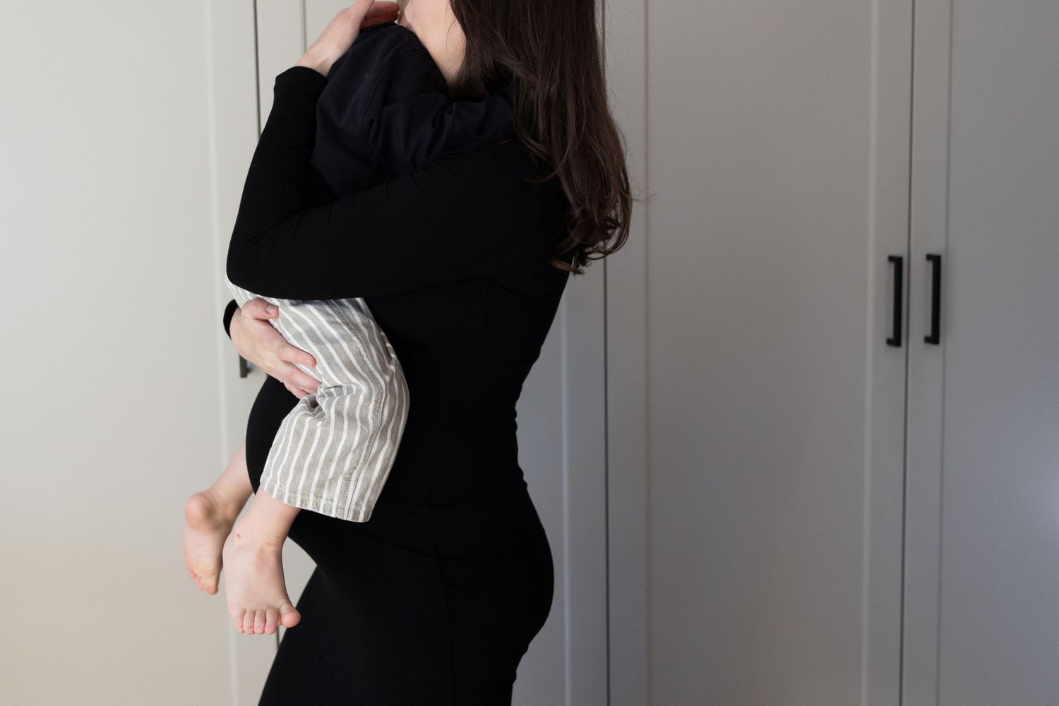 Woman in black dress holding a young child indoors near white wardrobe closet.