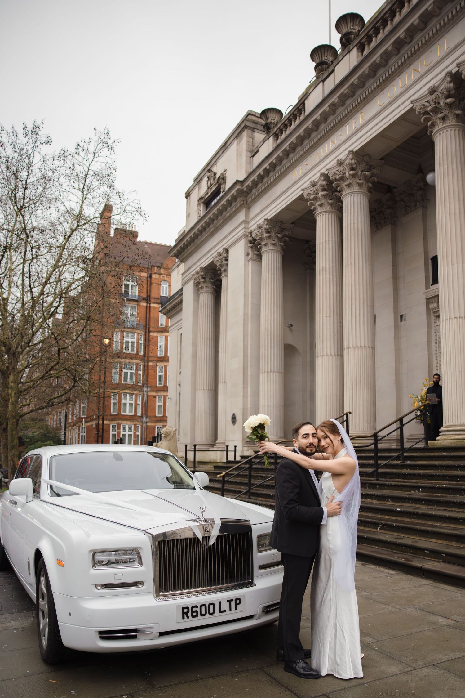 Bride and groom pose beside a white Rolls-Royce outside a grand neoclassical building with tall columns in London.