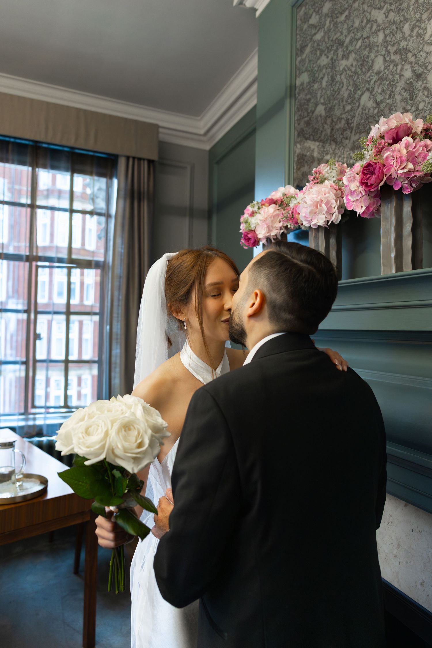 Bride holding white roses bouquet kissing groom near window with pink floral arrangement.
