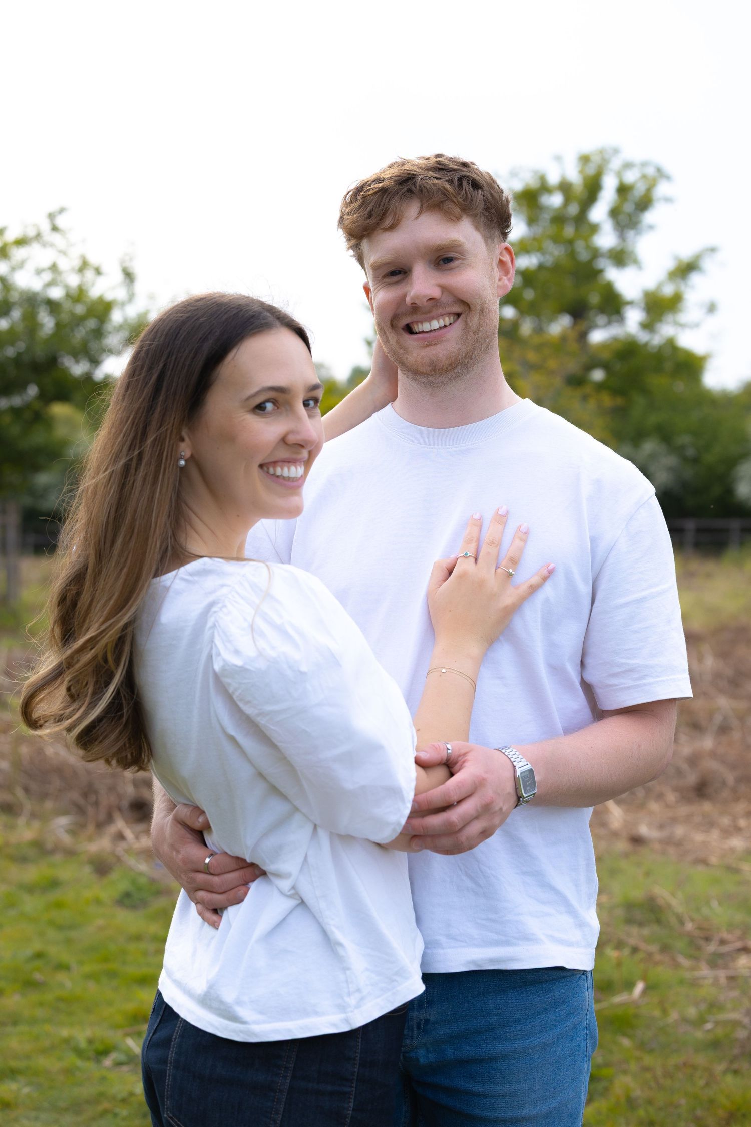 Smiling engaged couple in white shirts outdoors, woman displaying her engagement ring to the camera.
