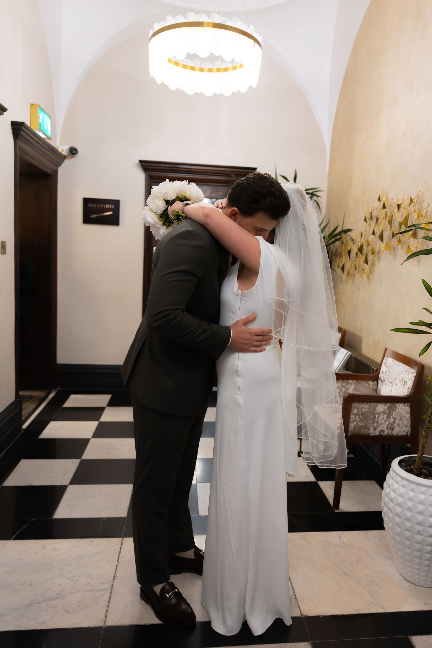 Newlyweds sharing first embrace in black and white tiled venue foyer after wedding ceremony.