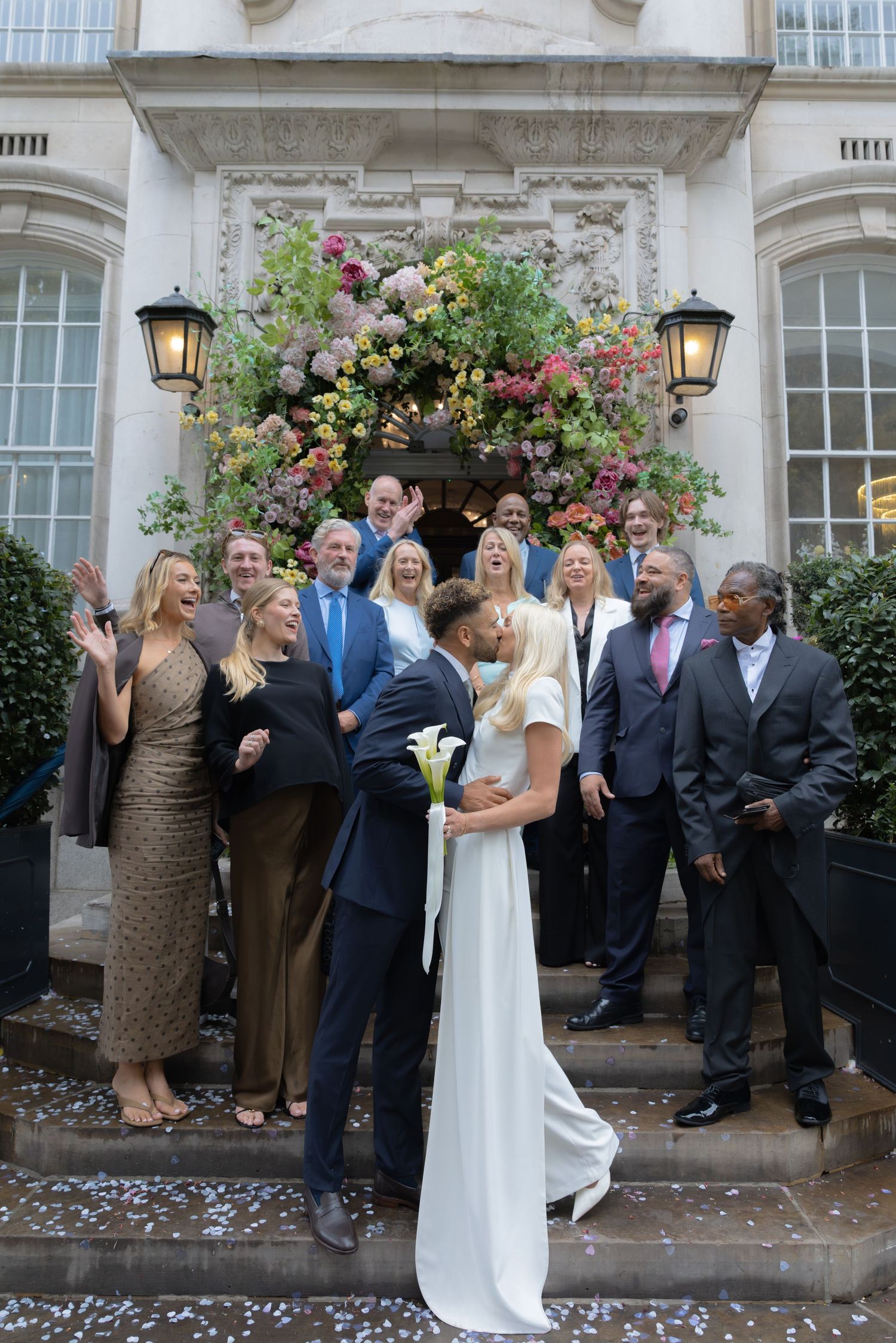 Bride and groom share a kiss on steps of elegant venue surrounded by cheering wedding guests and floral arch.