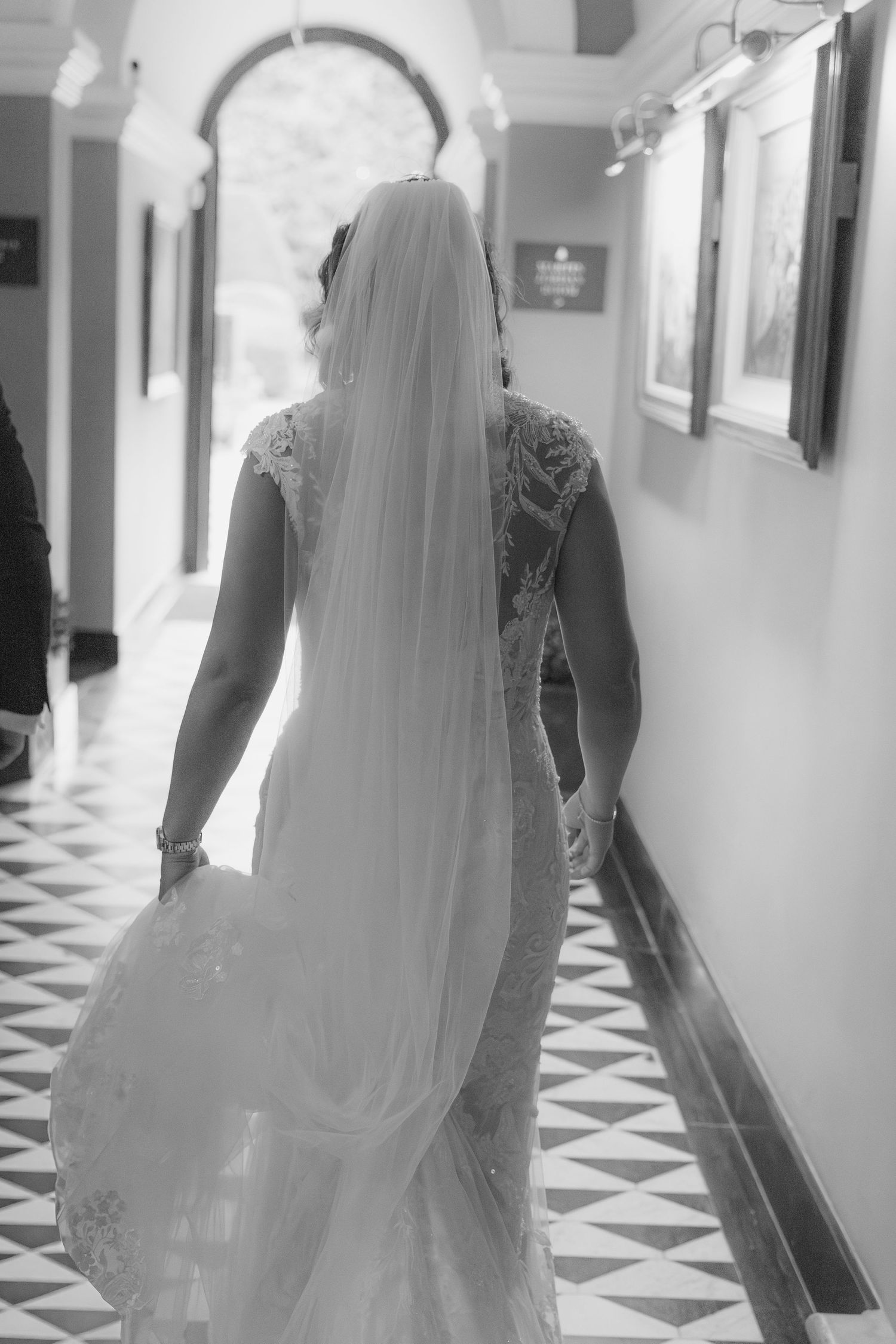 Elegant bridal gown with flowing veil captured in black and white along a geometric tiled hallway.
