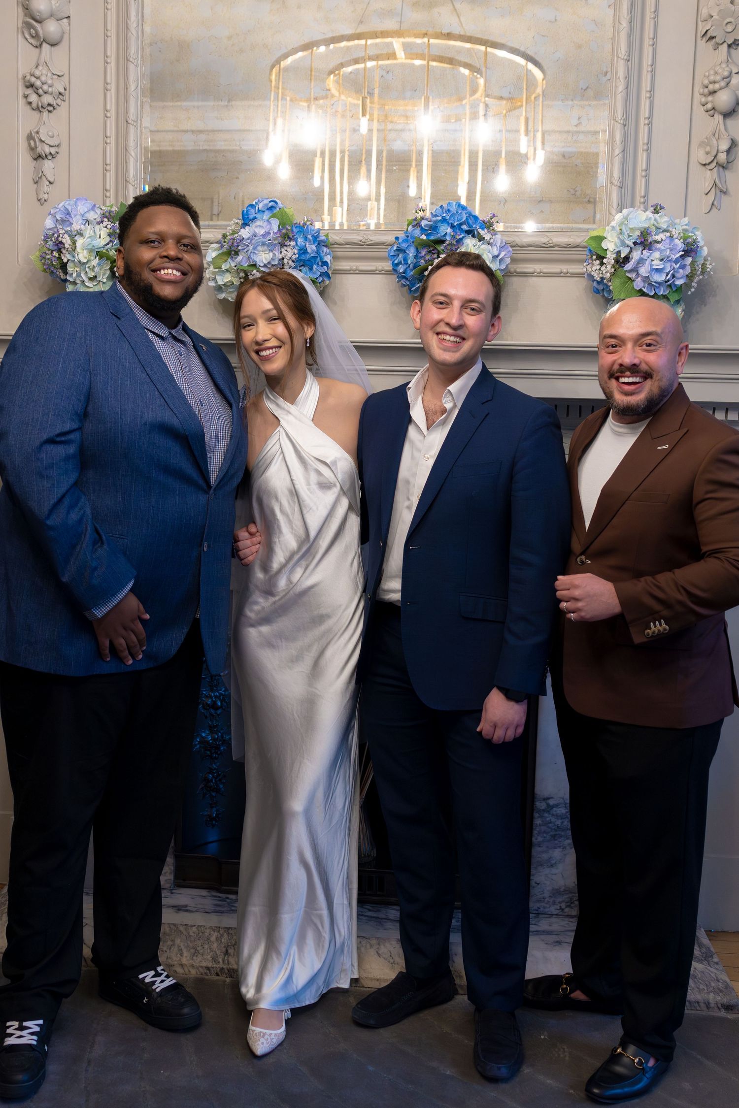 Bride and groom pose with two guests under blue hydrangea decorations at elegant wedding reception.
