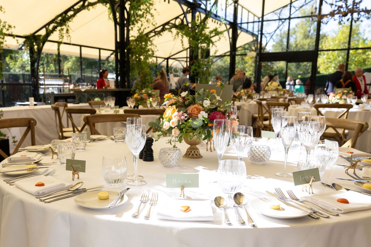 Elegant wedding reception table setting with white linens, wine glasses and floral centerpieces under a greenhouse-style venue.