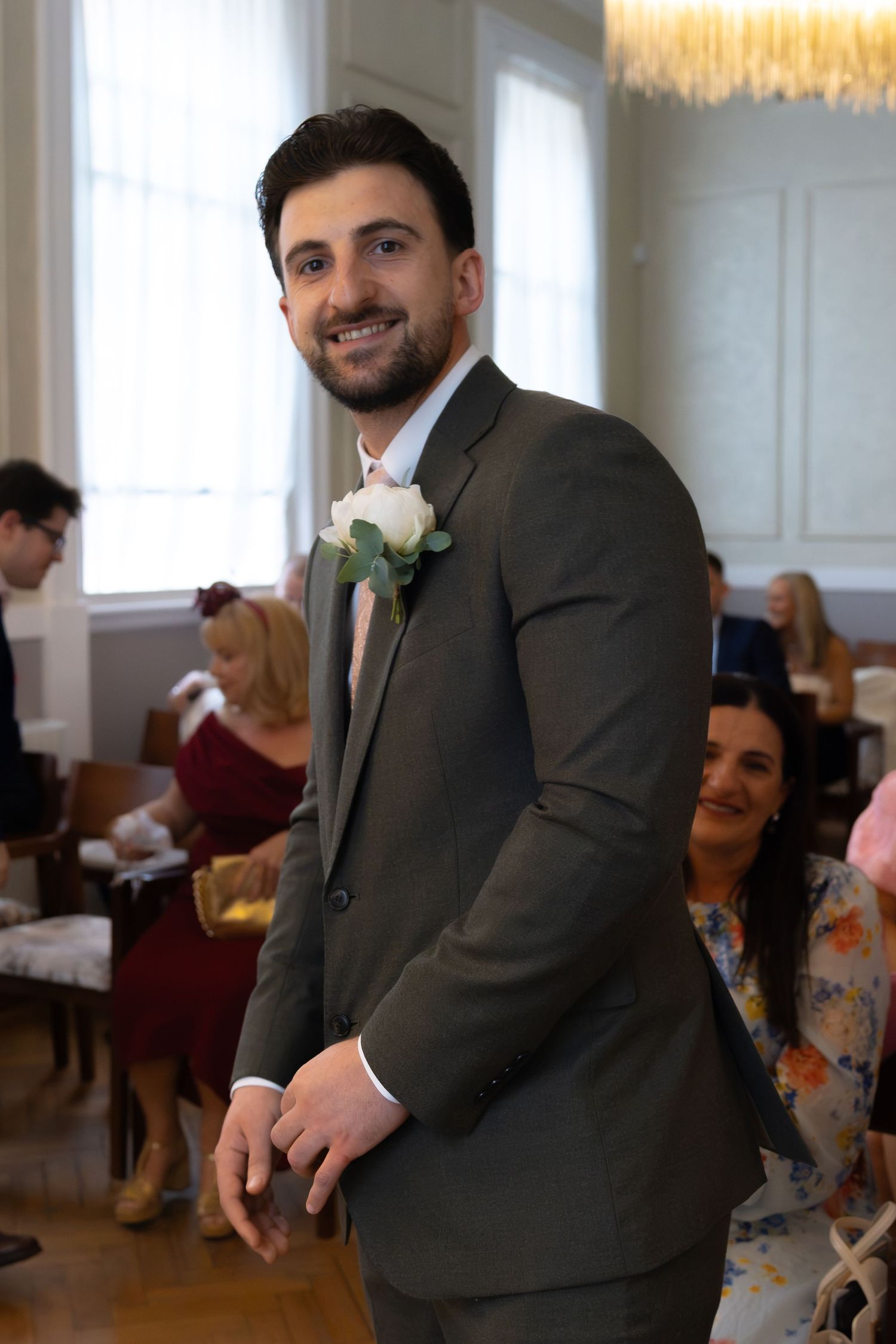 Smiling groom in gray suit with white boutonniere stands at wedding ceremony with seated guests behind.