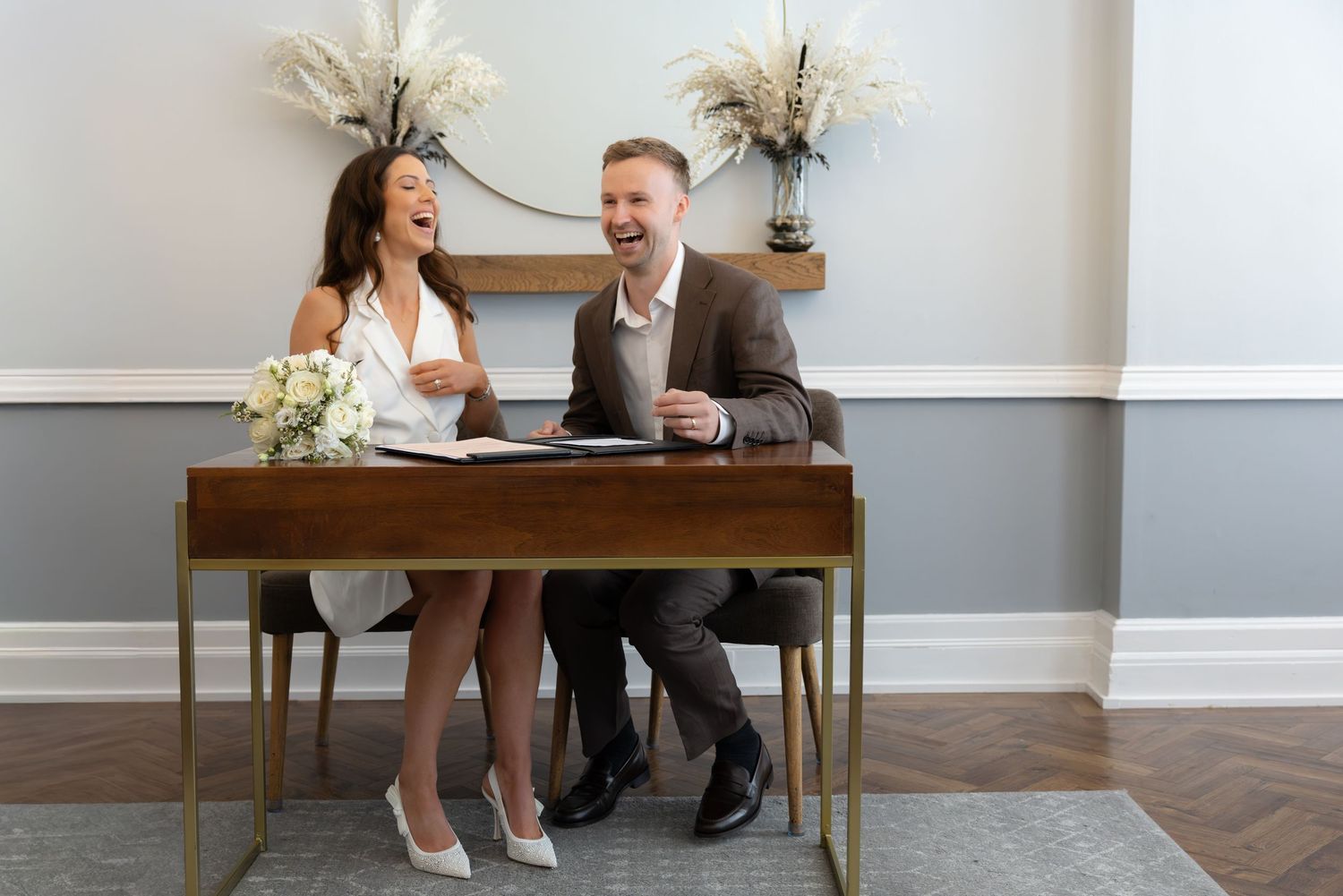 Happy couple laughing and signing marriage documents at a wooden desk in an elegant ceremony room.