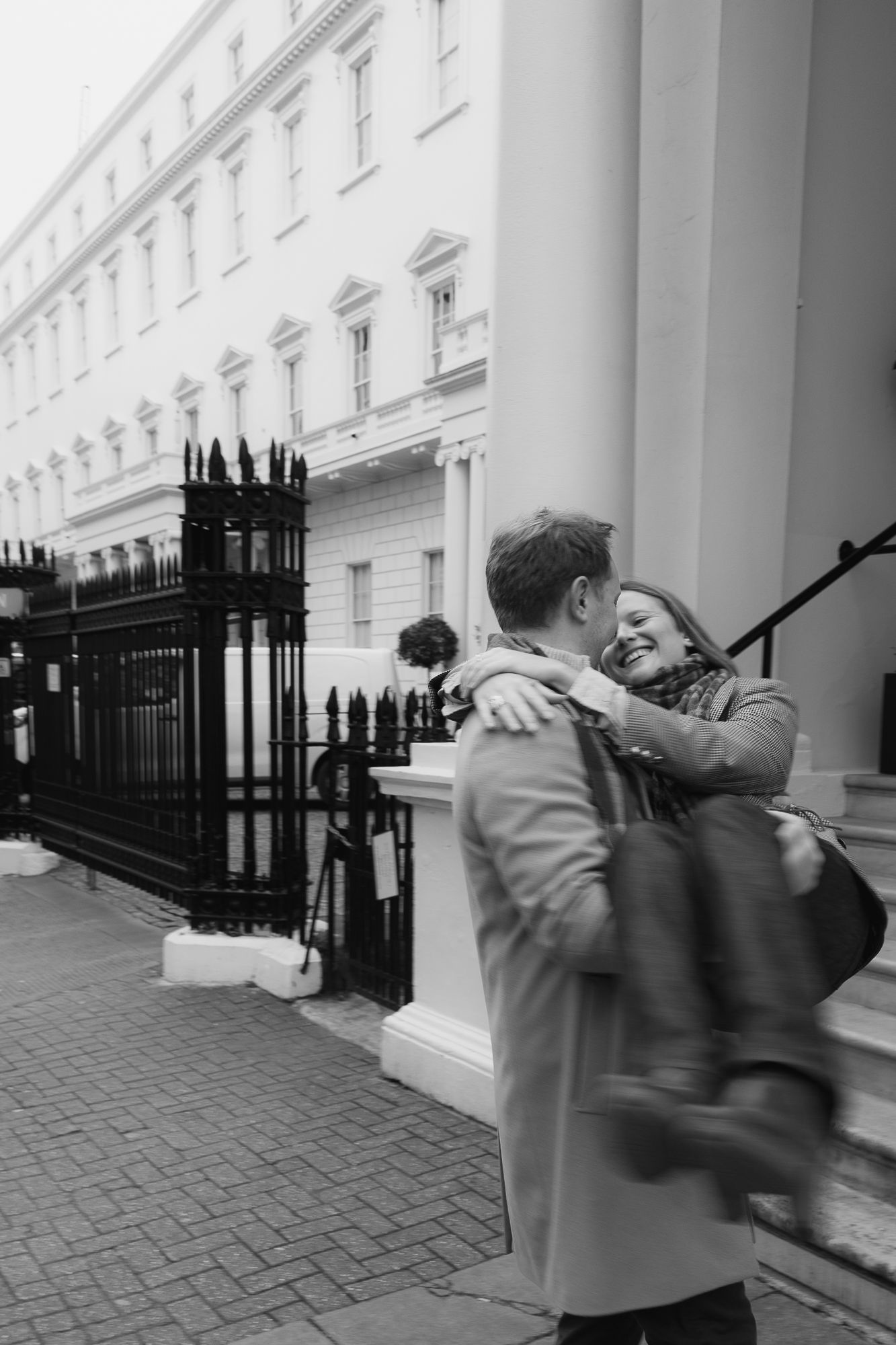 A couple shares an embrace on a cobblestone sidewalk next to an iron fence.