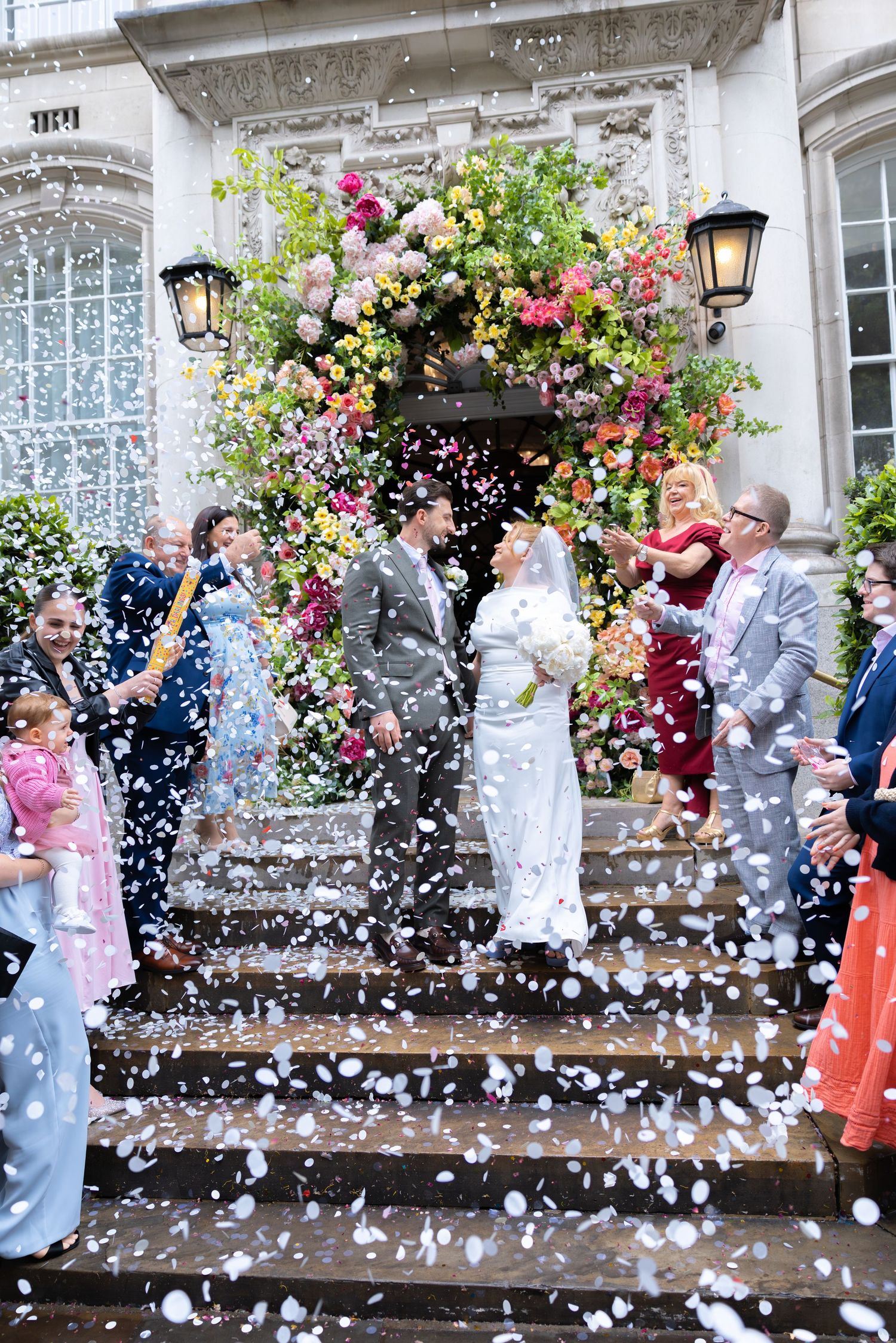 Bride and groom exit venue on steps showered in white confetti with colorful floral arch backdrop and cheering wedding guests.