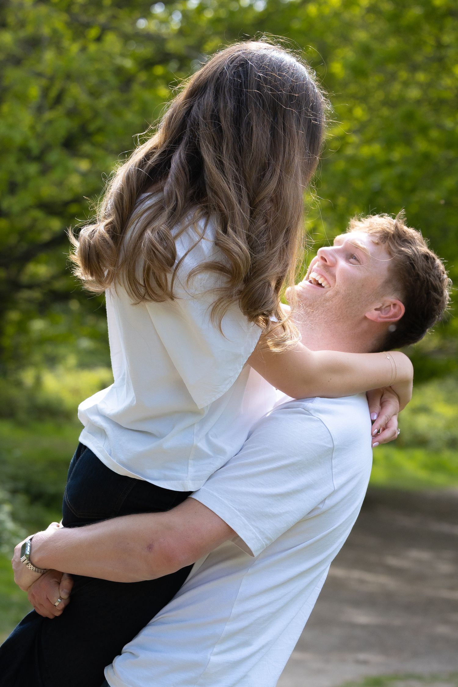 Happy young man lifting his girlfriend in a sunny green park, both wearing white tops.