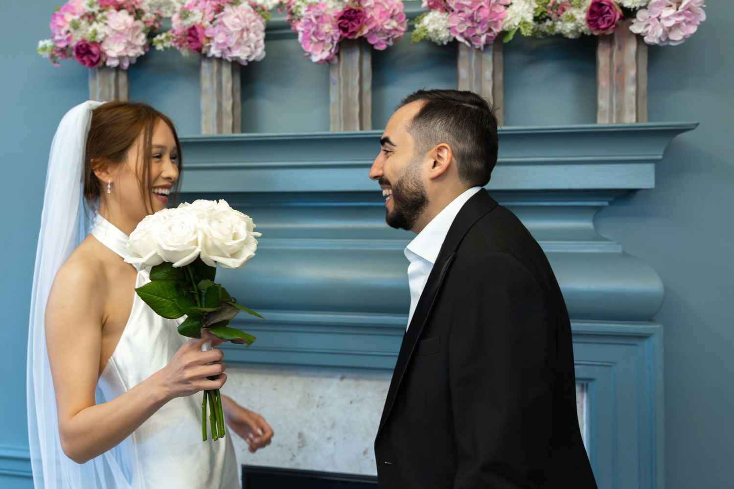 Bride holding white roses smiling at groom before blue fireplace with pink floral arch.