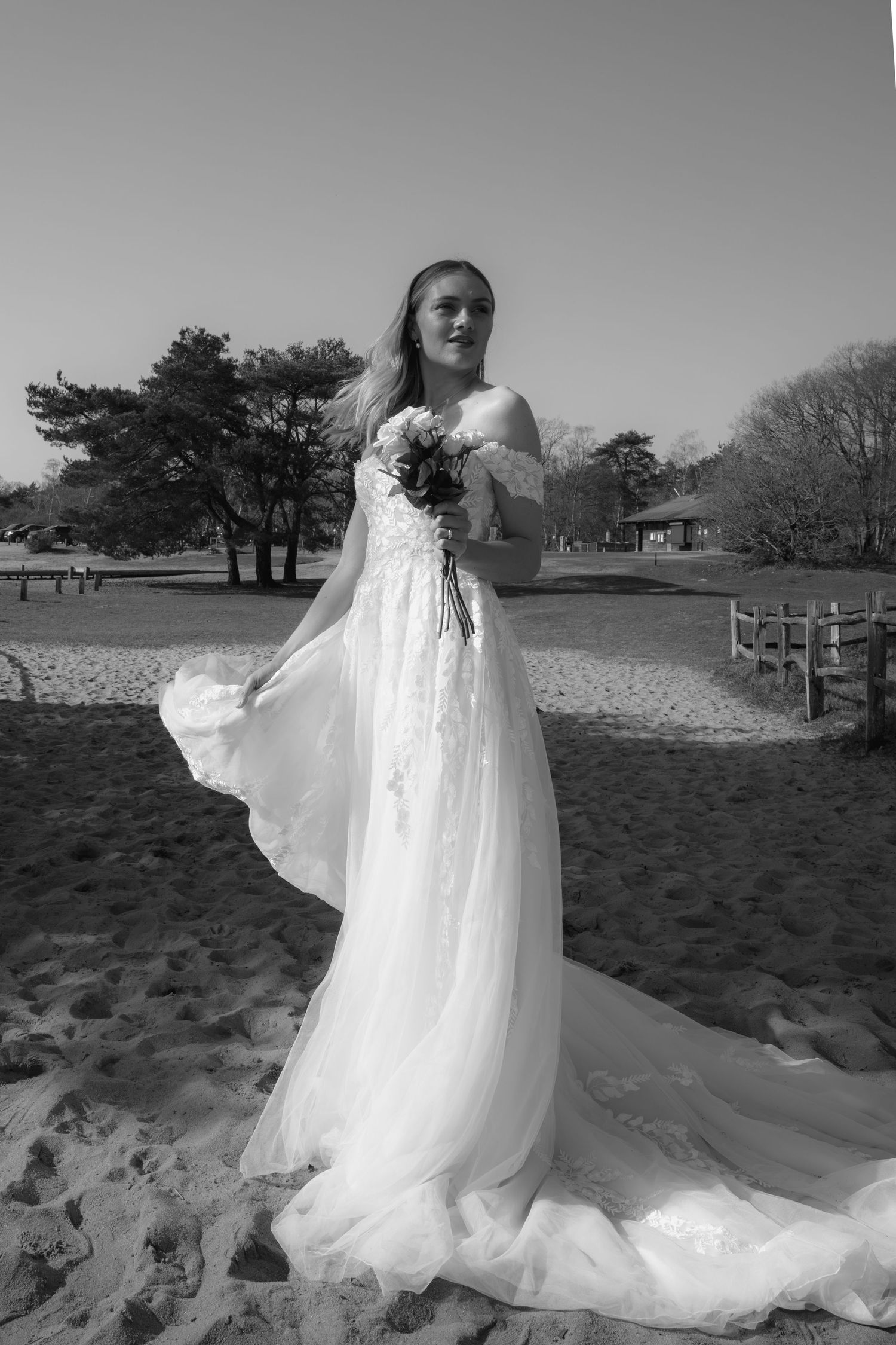Bride in flowing white wedding gown holding bouquet stands on sandy beach with trees in black and white photo.