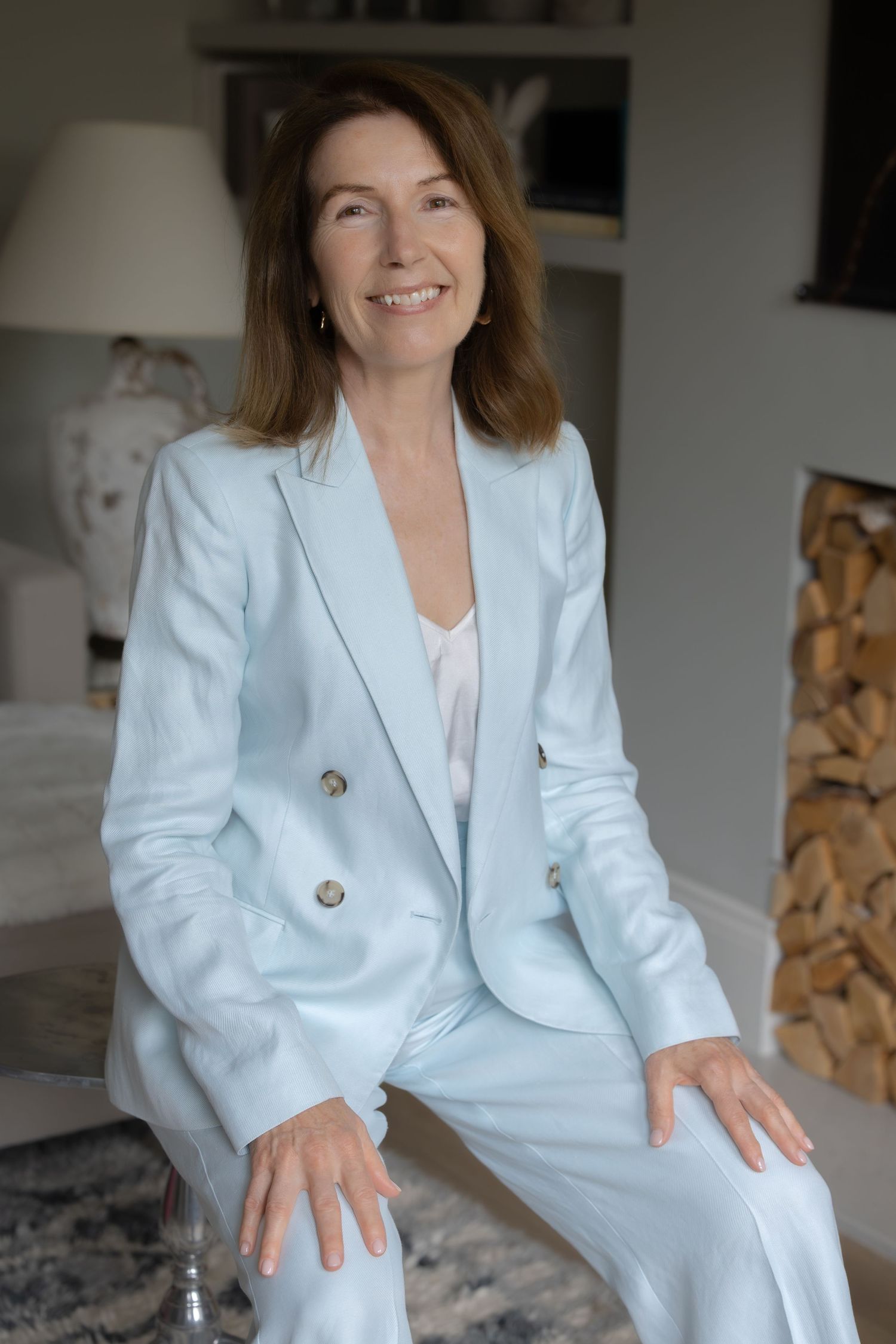 Smiling middle-aged woman in a light blue pantsuit sitting relaxed in a bright modern living room.