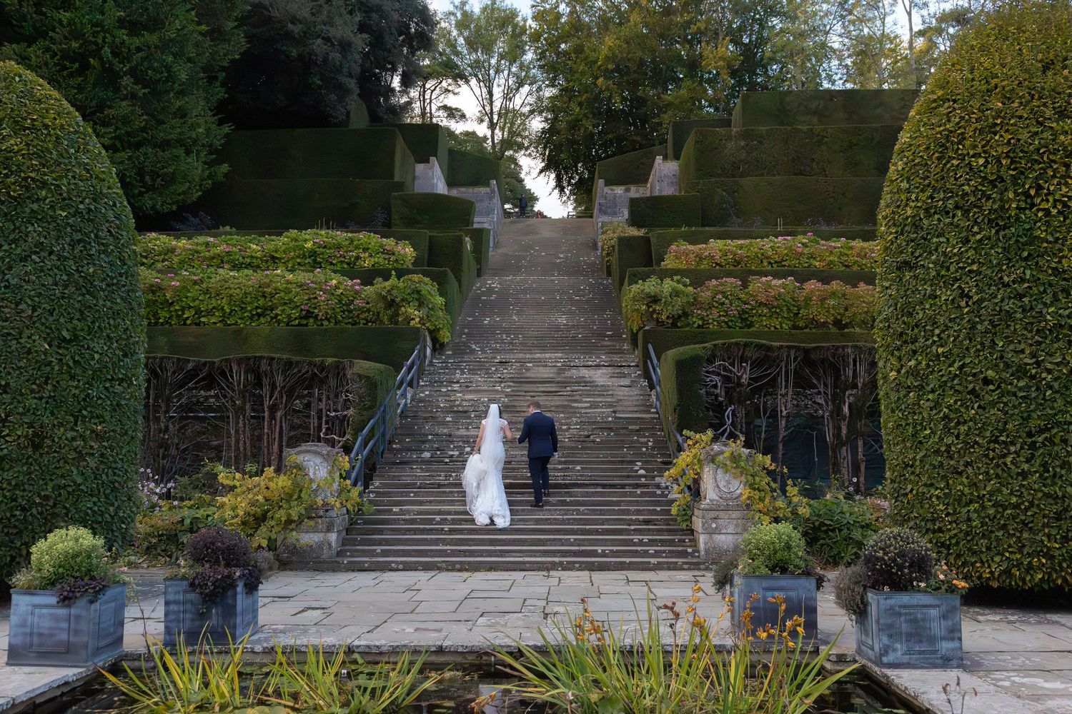 A stone stairway leads up through a formal garden with manicured hedges and trees creating a scenic walkway.