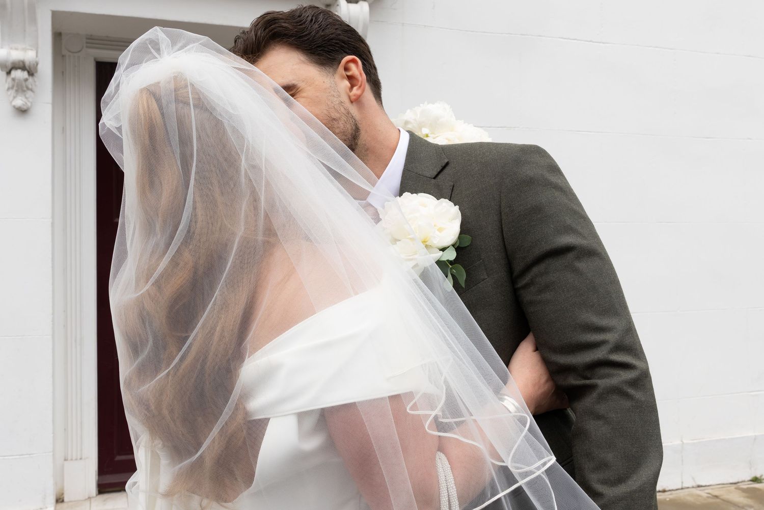 Bride and groom sharing a romantic kiss under a white veil on their wedding day outside a white building.