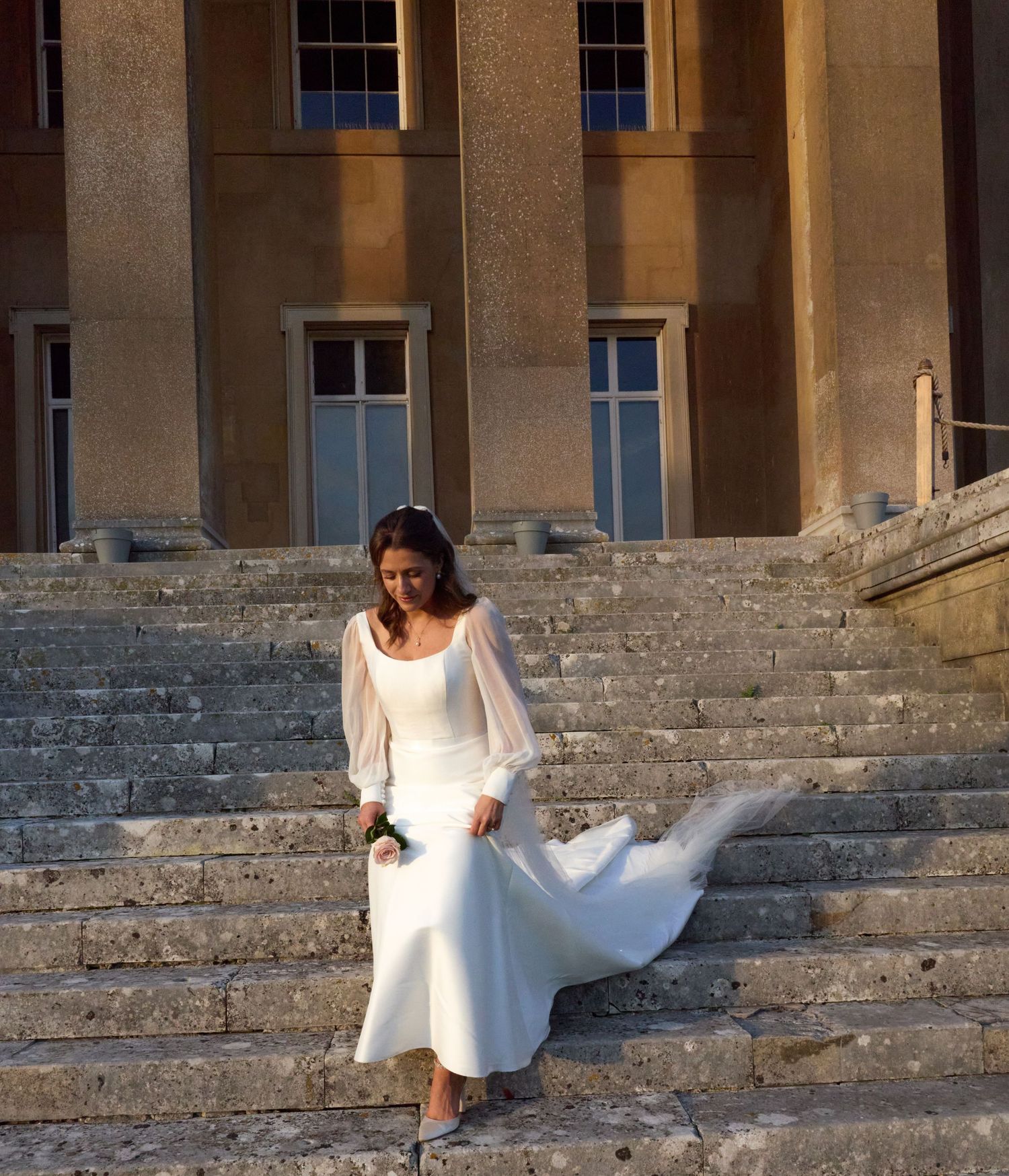 A bride in a flowing white dress sits on stone steps of a historic building at sunset.