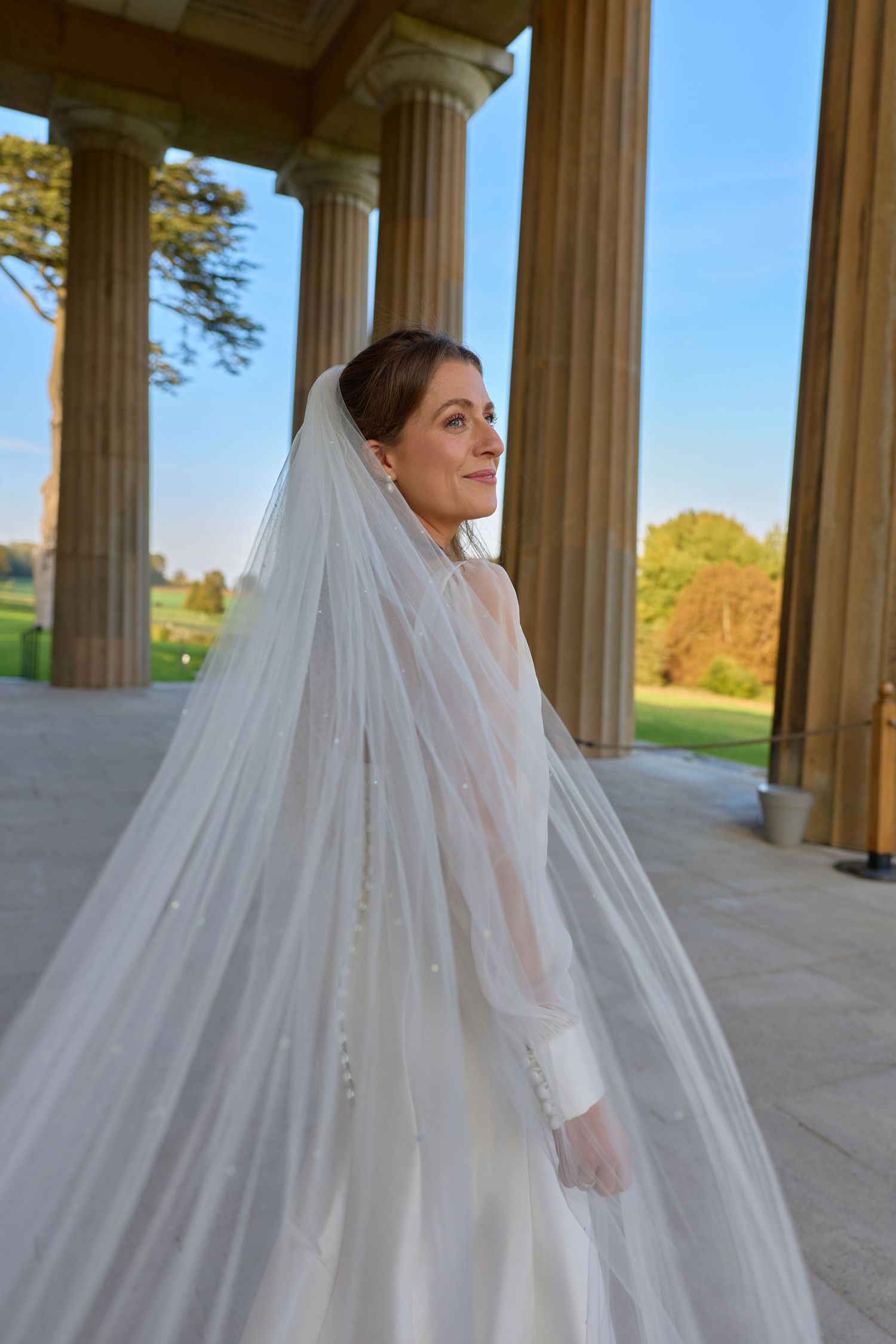 Elegant bridal portrait with flowing veil captured between majestic stone columns at golden hour.