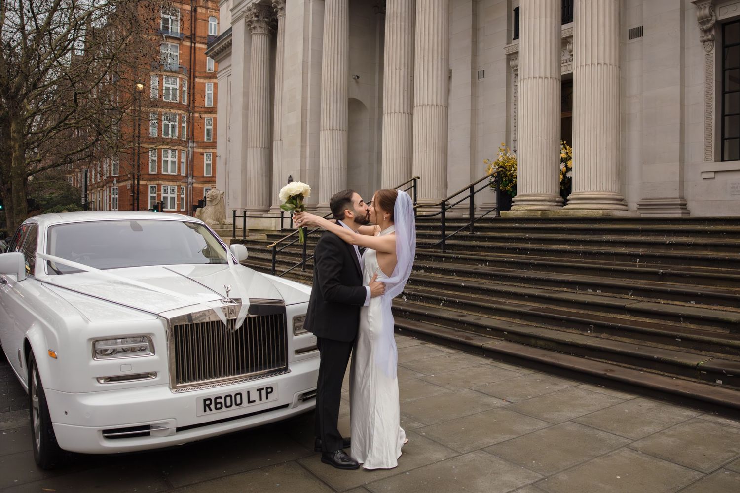Bride and groom sharing a kiss beside a white Rolls-Royce outside a grand building on their wedding day.