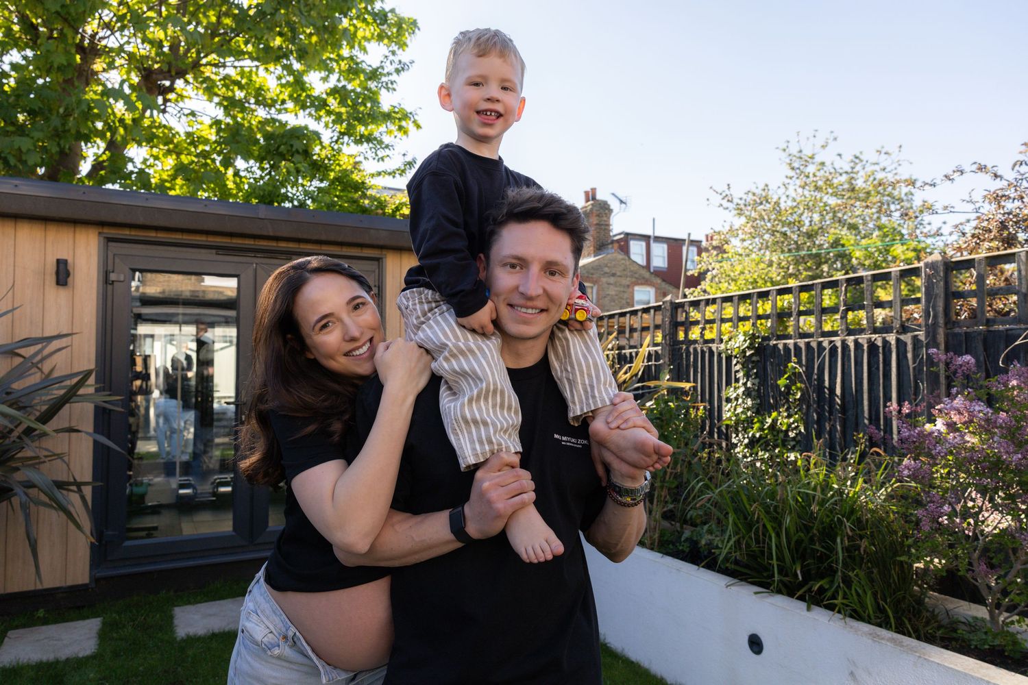 Pregnant woman and man with young boy on shoulders smiling in sunny garden.