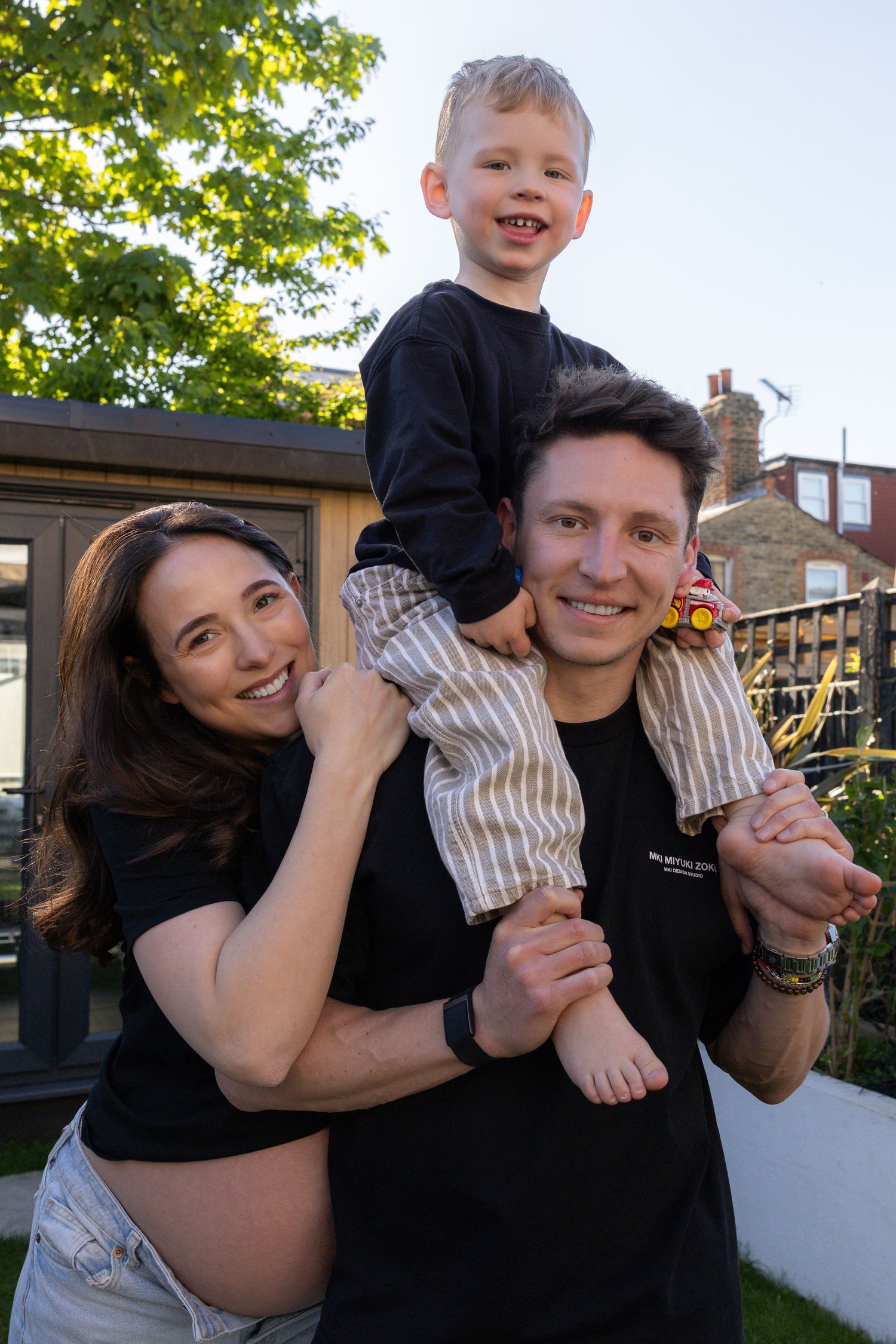 Happy family outdoors, man carrying young boy on shoulders while pregnant woman embraces them.