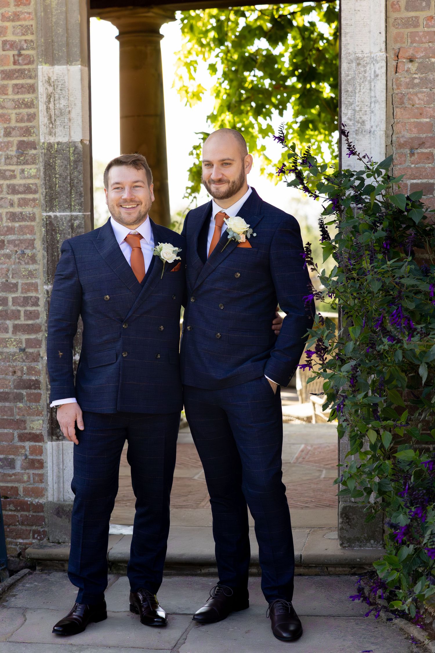 A series of groomsmen in navy suits and orange ties posing together under a brick archway.