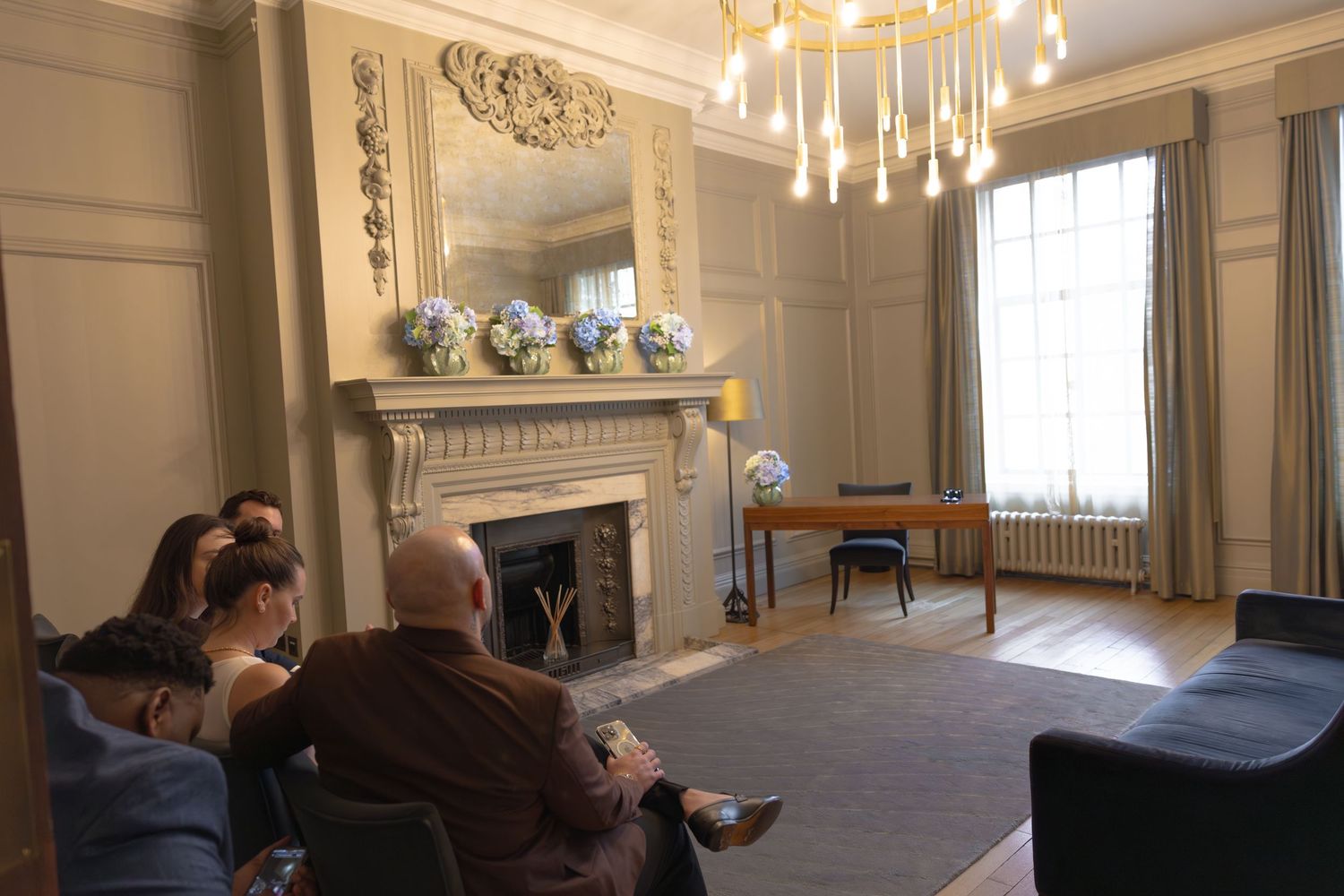Elegant living room with ornate fireplace, chandelier, and guests seated on sofas in a classic interior.