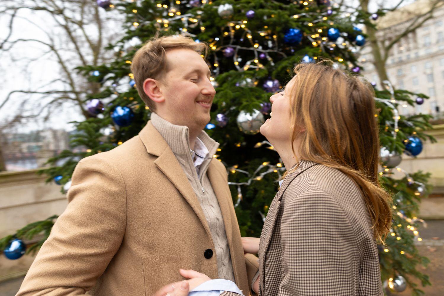 Two people share a joyful moment in front of a Christmas tree decorated with blue ornaments.