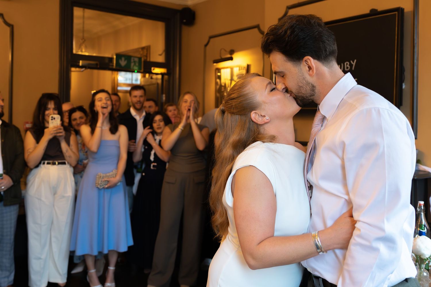Couple shares first kiss at wedding reception as guests applaud in elegant venue.
