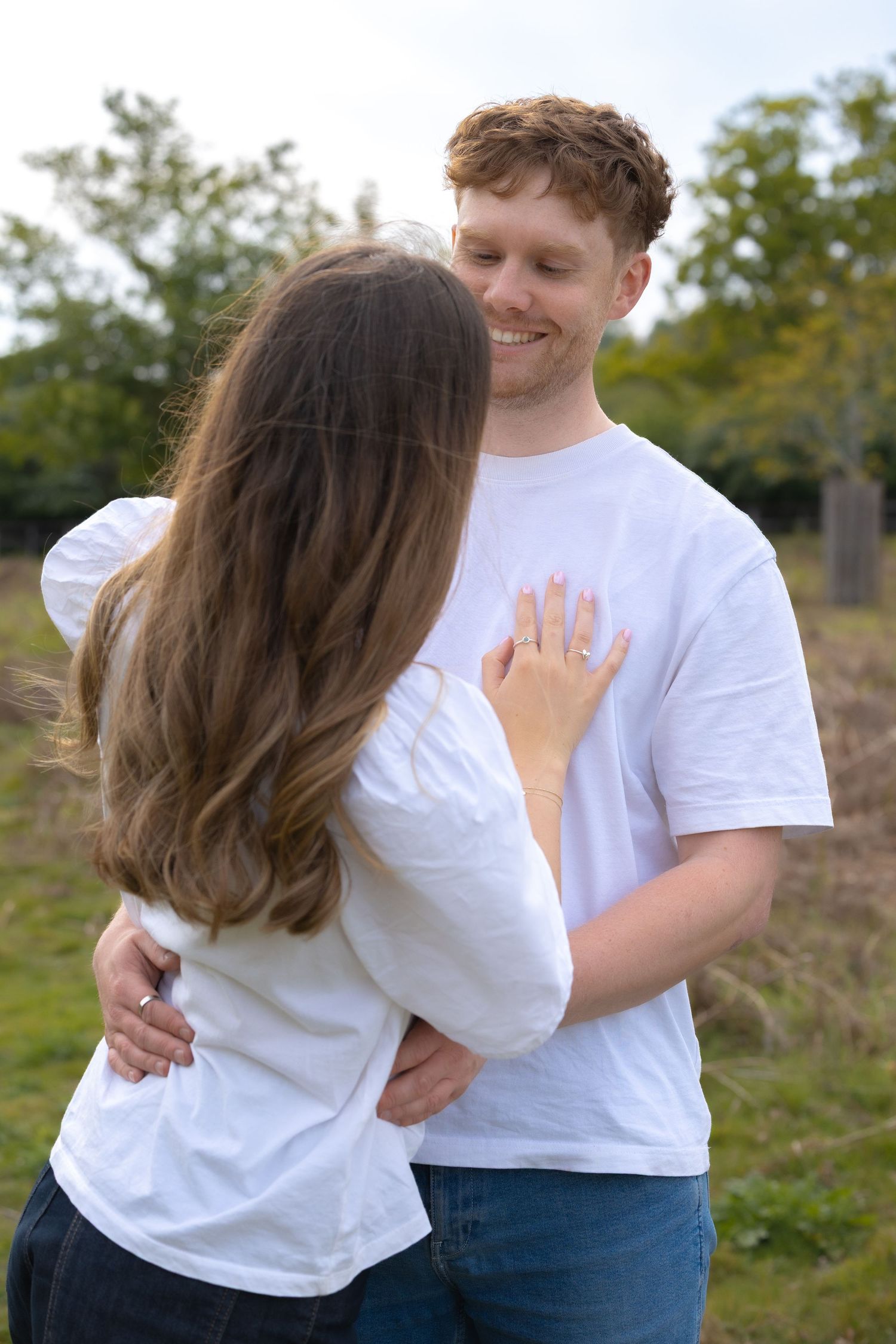 Smiling redhead man embracing woman outdoors, she displays an engagement ring on her hand.