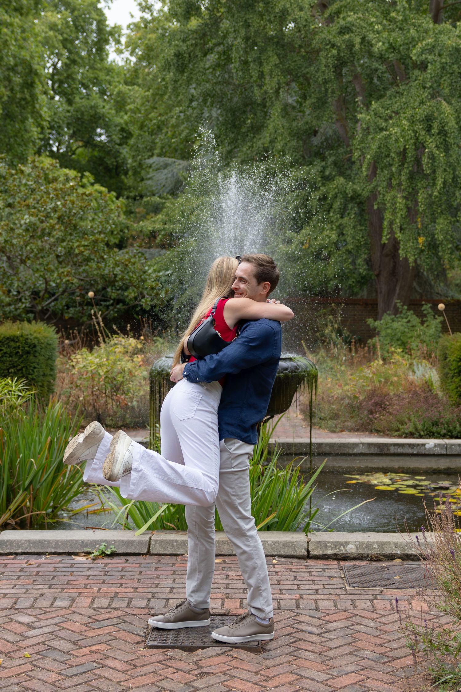 Couple embracing and kissing by a garden fountain on brick path, surrounded by lush green trees.