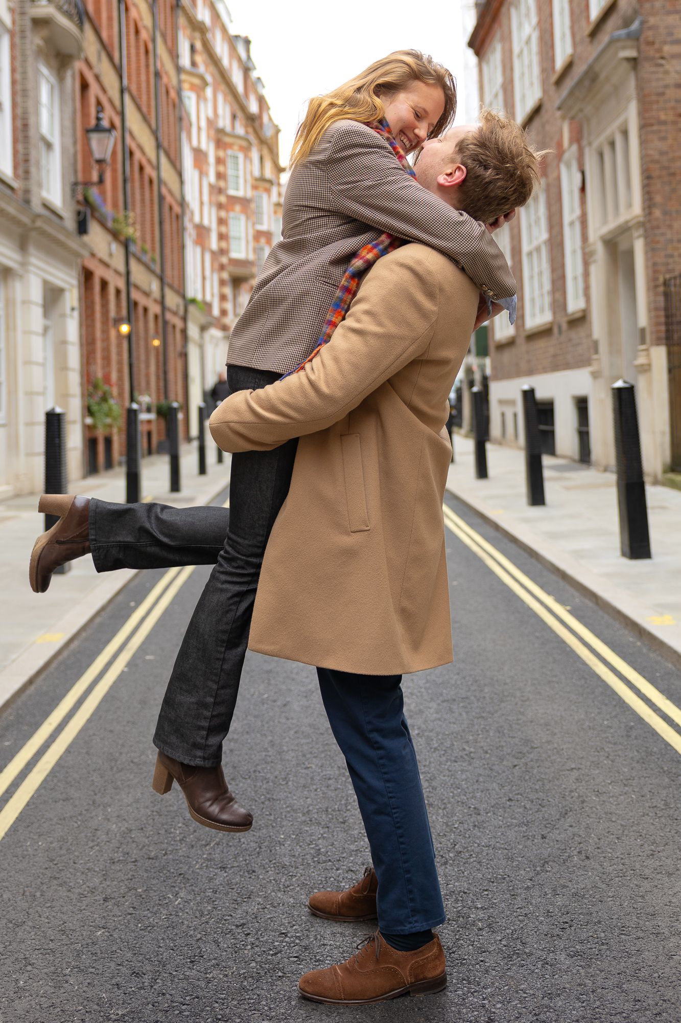 A playful couple embraces on a cobblestone street lined with brick buildings.