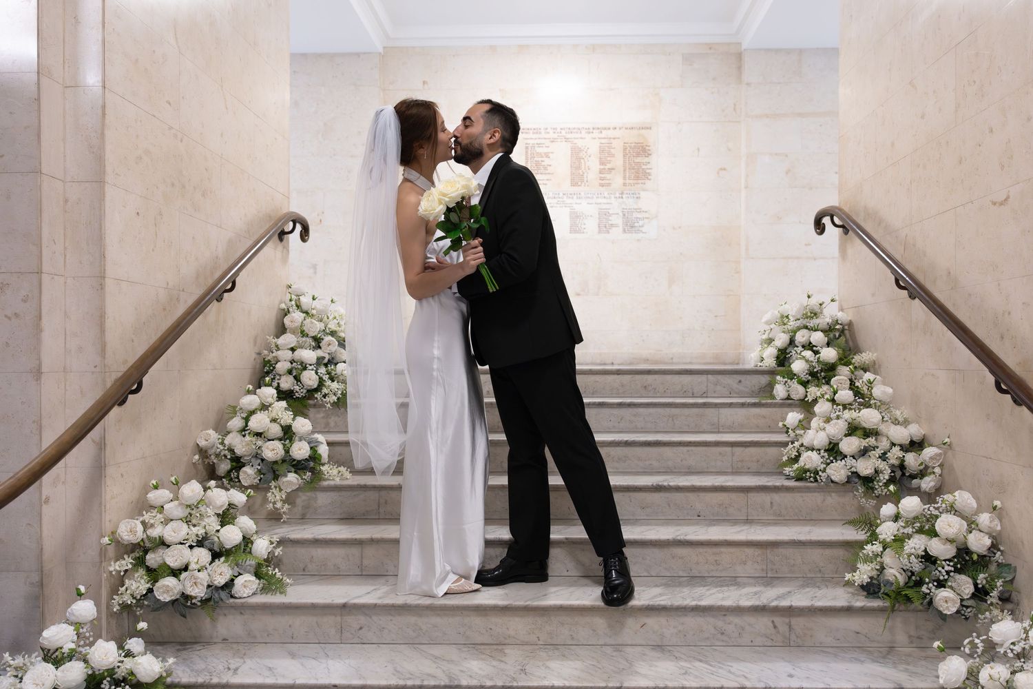 Bride and groom share a kiss on flower-adorned marble staircase, bride holding white bouquet in elegant venue.
