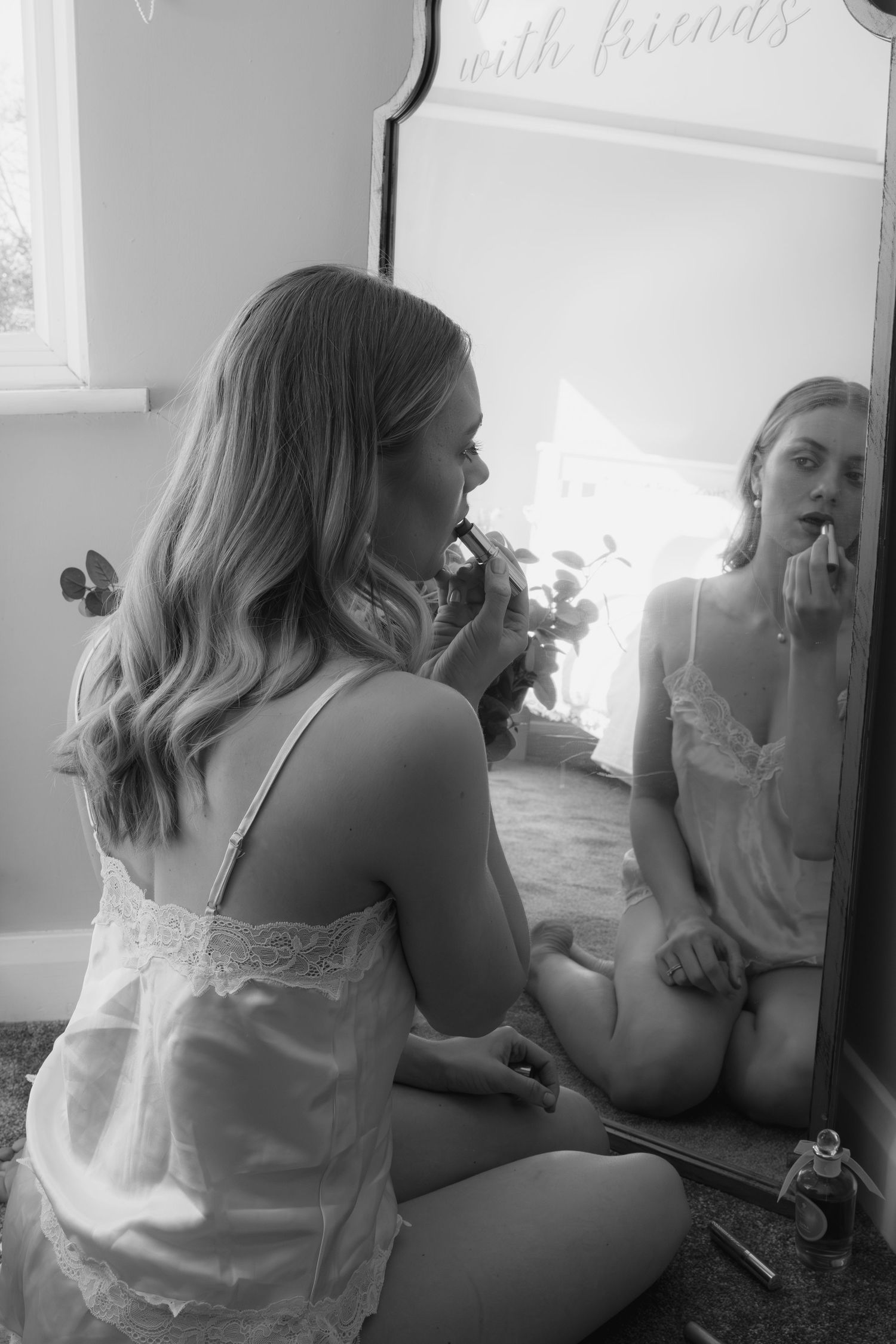 Young woman in white slip applies lipstick while sitting before large mirror in black and white photo.