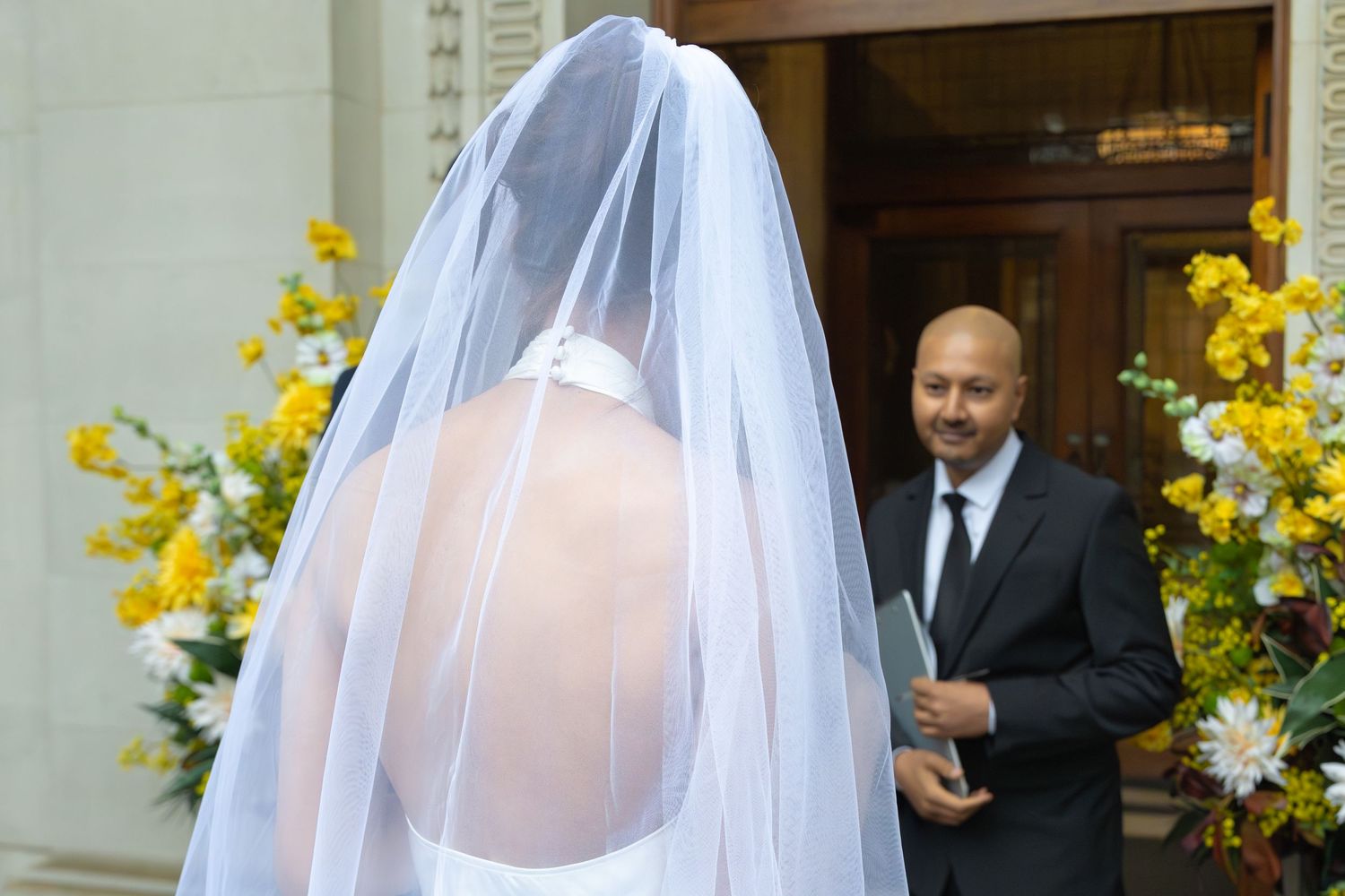 Bride in white gown and veil stands before groom in black suit at elegant wedding ceremony entrance.