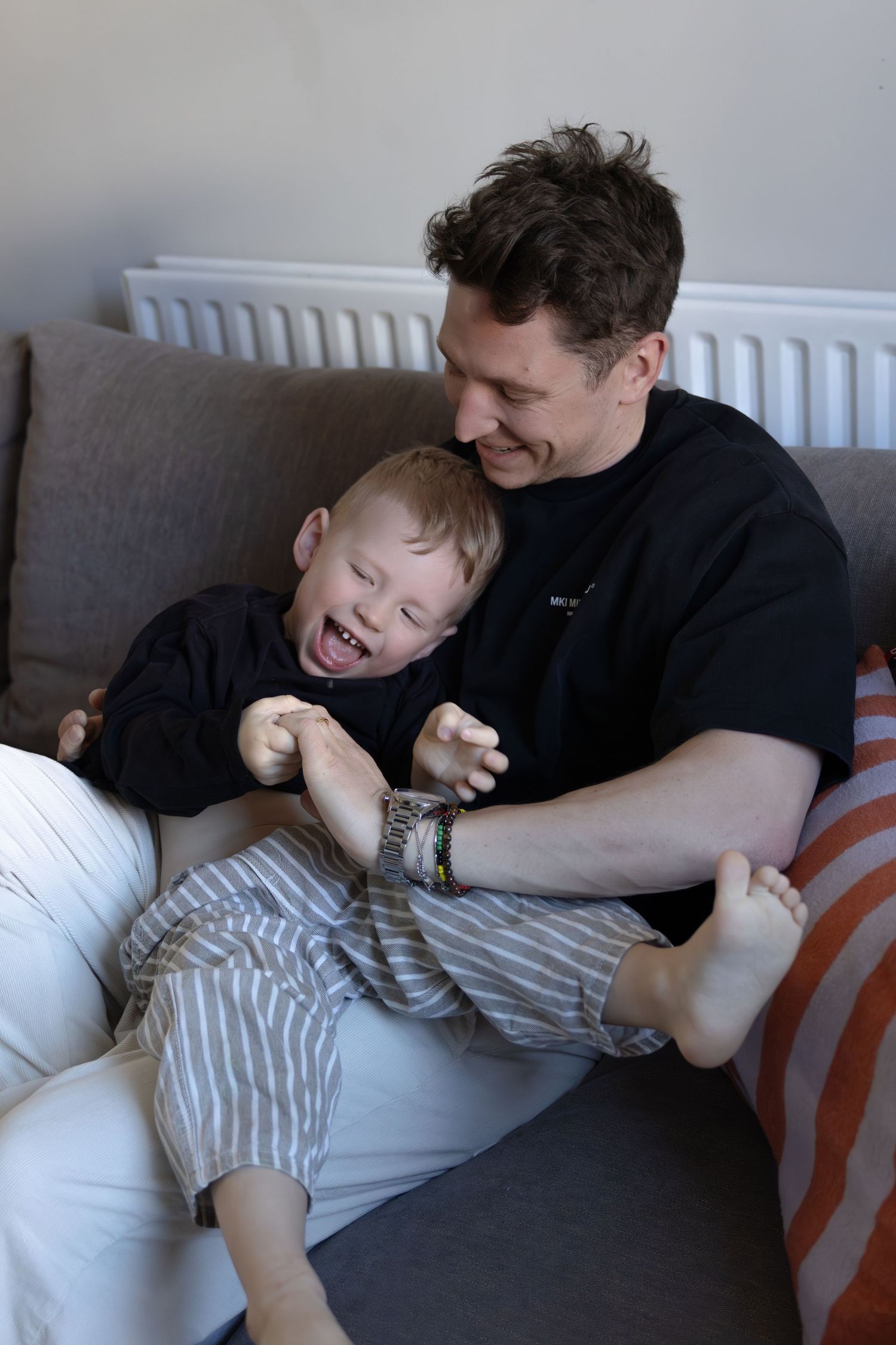 Father laughing while playfully tickling happy toddler son on living room sofa.